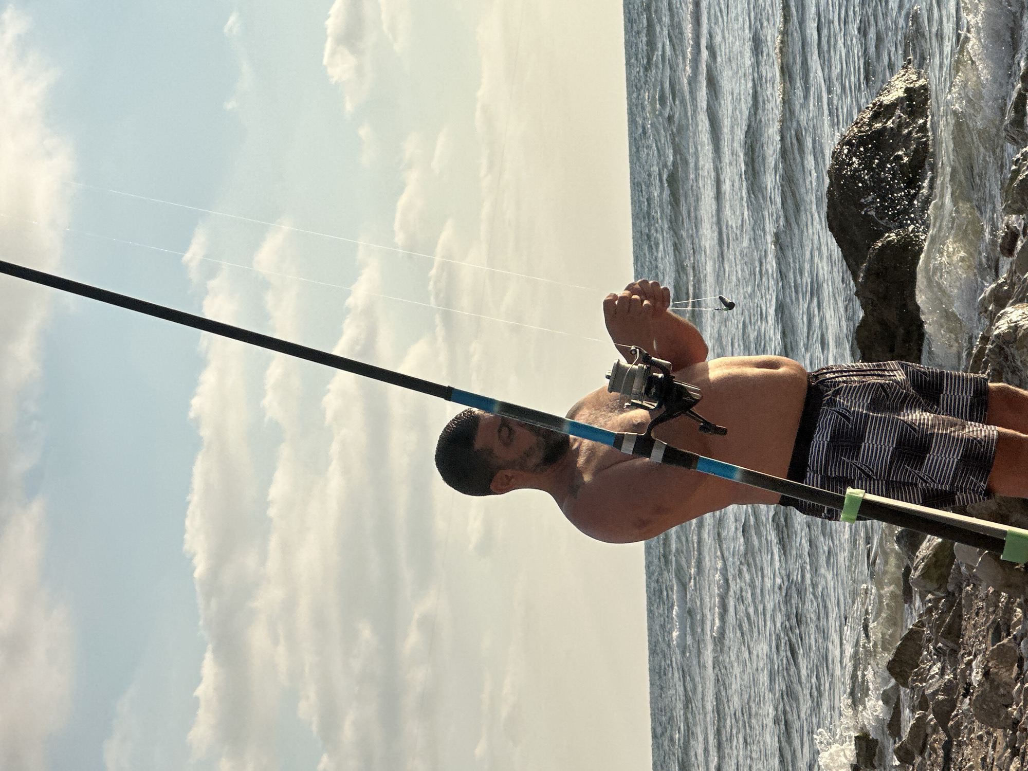 A man is fishing on a rocky shoreline at the sea. The man is wearing patterned swim trunks and no shirt. He is holding a fishing rod with a reel and appears to be adjusting the fishing line. The sea is wavy, and there are rocks and a large boulder in the foreground. The sky is partly cloudy with a light yellow hue, suggesting it is either sunrise or sunset. The image is likely taken in Haifa, Israel, given the presence of the sea and the characteristic rocky coast.