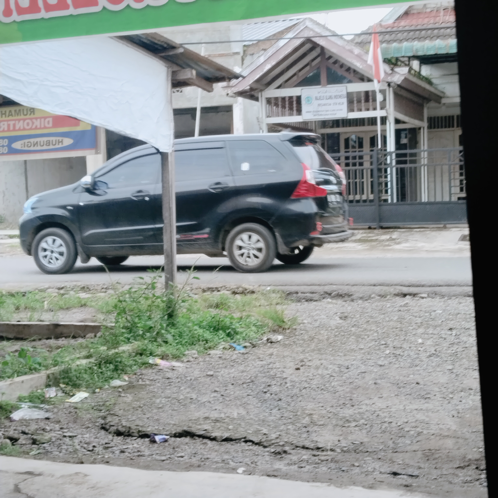In Talun Kenas, Indonesia, a black minivan is parked on a paved road beside a property with a white sign and a flag. The vehicle is positioned to the left of the frame, facing forward, with its passenger side visible. A white tarp hangs over the front of the property, partially obscuring a banner with text. The ground in front of the property is covered with gravel and patches of green vegetation. A cracked concrete barrier runs along the edge of the road. The background shows a building with a tiled roof and a fence. The scene appears to be a street view in a residential or commercial area.