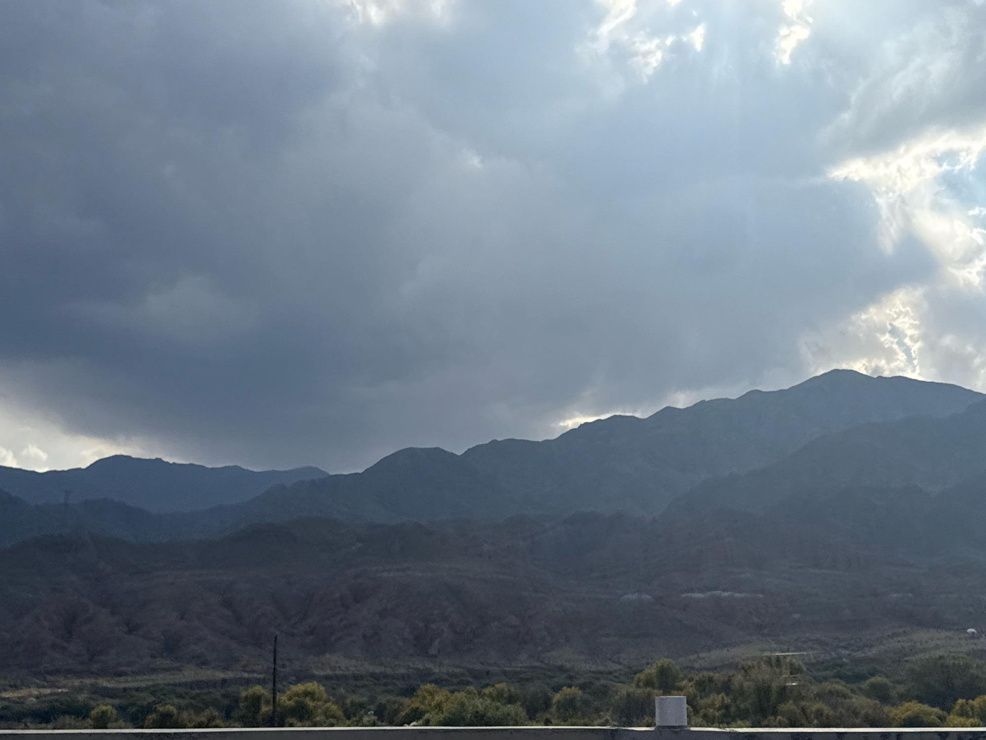The image depicts a mountainous landscape under a dramatic sky filled with dark, heavy clouds. The mountains are layered, with the closest peaks appearing darker and more detailed, while the furthest ones fade into a muted blue. The terrain in front of the mountains is dry and sparse, with some scattered vegetation and evidence of erosion. A subtle hint of human presence is visible in the form of a light-colored post or structure on the right side of the foreground, and a thin vertical pole extending from the mid-ground. The lighting suggests either early morning or late afternoon, with sunlight breaking through the clouds in places, creating highlights on the mountain ridges and the sky. The overall impression is one of rugged natural beauty and a sense of impending weather. The location is not identifiable from the image itself as being in the city of Unknown, Kyrgyzstan.