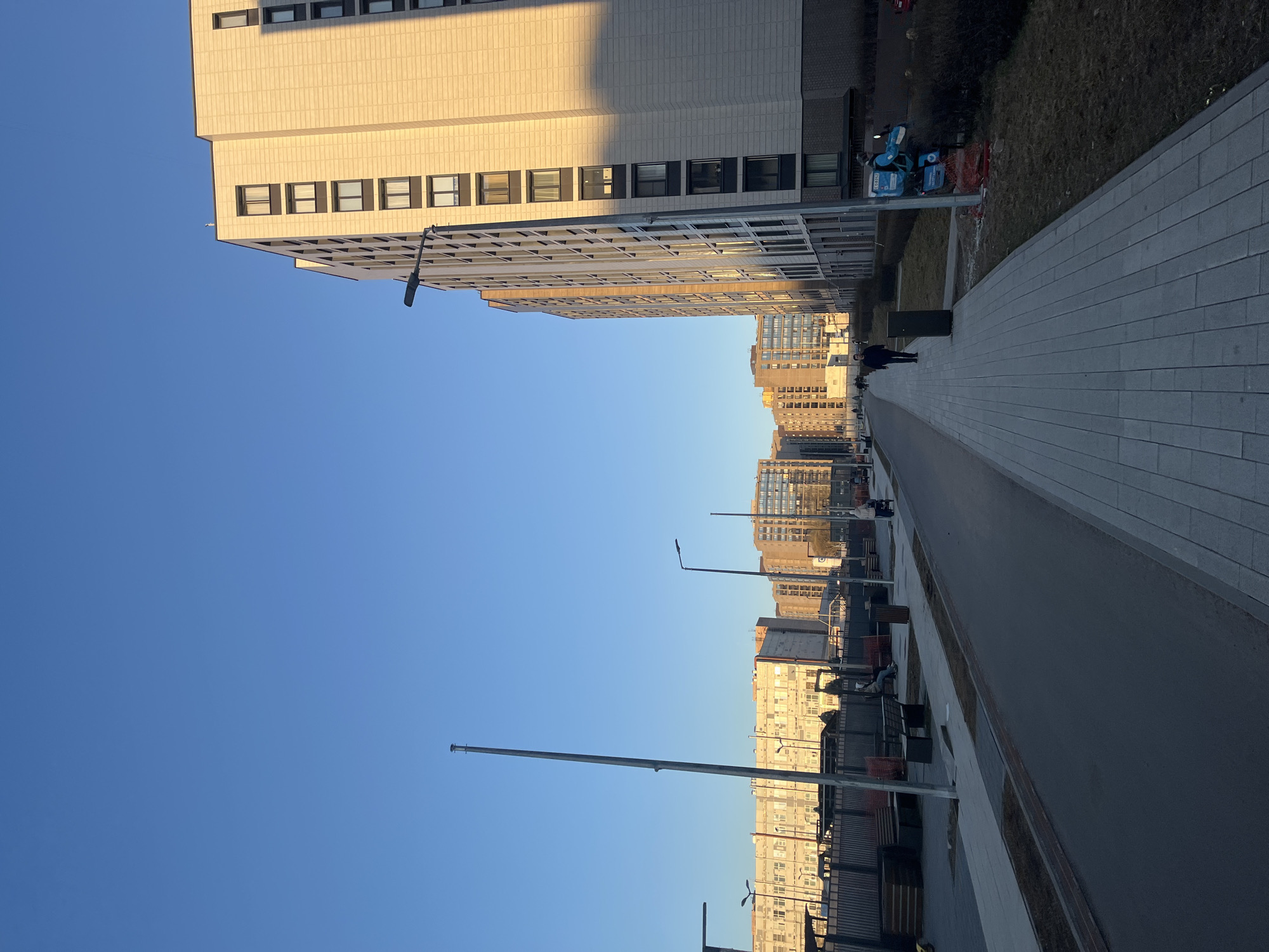 The image depicts a street in Saint Petersburg, Russia, lined with modern apartment buildings. The sky is clear blue. The buildings are light-colored and have a repetitive pattern of windows. A wide paved walkway runs along the right side of the street, with some people walking on it. Several streetlights are visible along the walkway. There are also some benches. The image is taken from a low angle, giving a perspective of the buildings towering over the street.