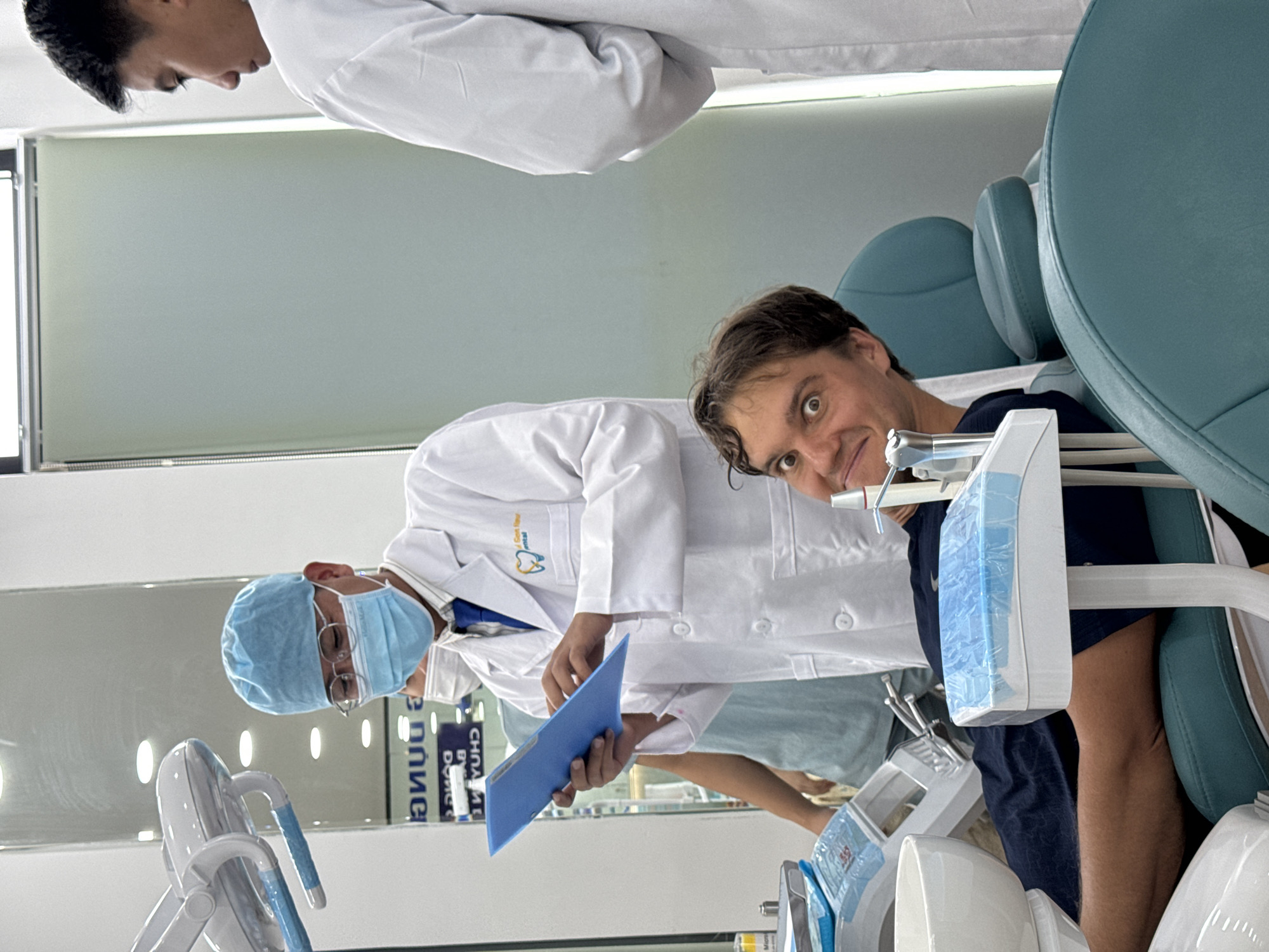 The image, when rotated to its correct orientation, captures a brightly lit scene inside a dental clinic. A male patient with dark, slightly curly hair is seated in a teal-colored dental chair in the foreground, looking directly at the camera with a wide-eyed, playful or surprised expression. He is wearing dark blue clothing.
Standing next to the patient is a male dental professional, possibly a dentist or assistant, wearing a white lab coat, a light blue scrub cap, and a matching light blue surgical mask. He holds a blue folder or clipboard in his hands. A logo on his lab coat features a stylized tooth and appears to include the word "Dental" or "Smile Dental." Another individual in a white lab coat is partially visible in the upper part of the frame.
The setting is a modern and clean clinic with white walls and bright lighting. Various dental instruments and components of the dental unit are visible, some covered in blue protective plastic. In the background, on a reflective surface, reversed Vietnamese text can be seen, likely from a mirrored sign. Portions of the text, such as "NHA KHOA" (meaning "Dental Clinic"), are discernible, consistent with the location context of Phu Thuy Ward, Vietnam. The scene depicts a routine visit, with the patient seemingly enjoying a lighthearted moment despite the medical setting.