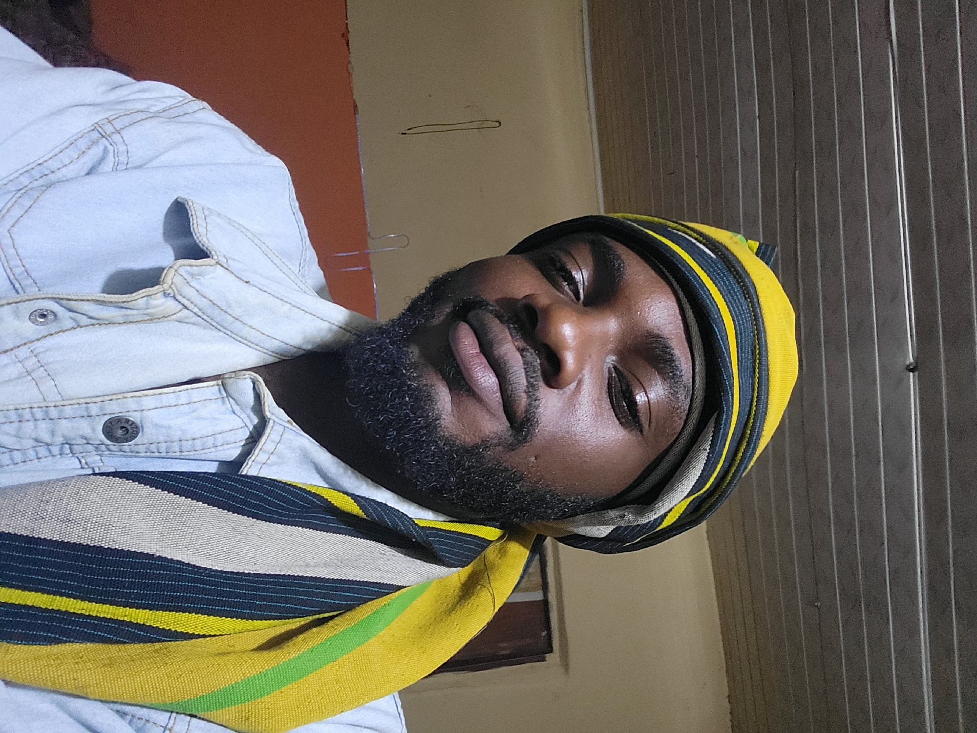 The image, rotated 90 degrees clockwise, presents a close-up portrait of a dark-skinned man, an adult with a full beard and mustache, looking directly at the camera with a neutral expression. He is wearing a light blue, denim-style shirt or jacket with visible stitching and metal buttons.
A multi-colored fabric wrap or scarf is draped around his head and neck/shoulder. This distinctive accessory features prominent horizontal stripes in yellow, dark blue/black, light gray/white, and a thin line of green.
The background suggests an indoor setting in Jos, Nigeria. To the left, a plain orange-brown wall is visible. To the right, there's a beige or light brown wall, on which a thin, dark, horizontal line is drawn, and a metal hook or hanger is present. Further to the right, a section of corrugated metal siding, possibly a wall or door, fills the frame. The lighting appears to be artificial and somewhat subdued. No legible text is visible in the image.