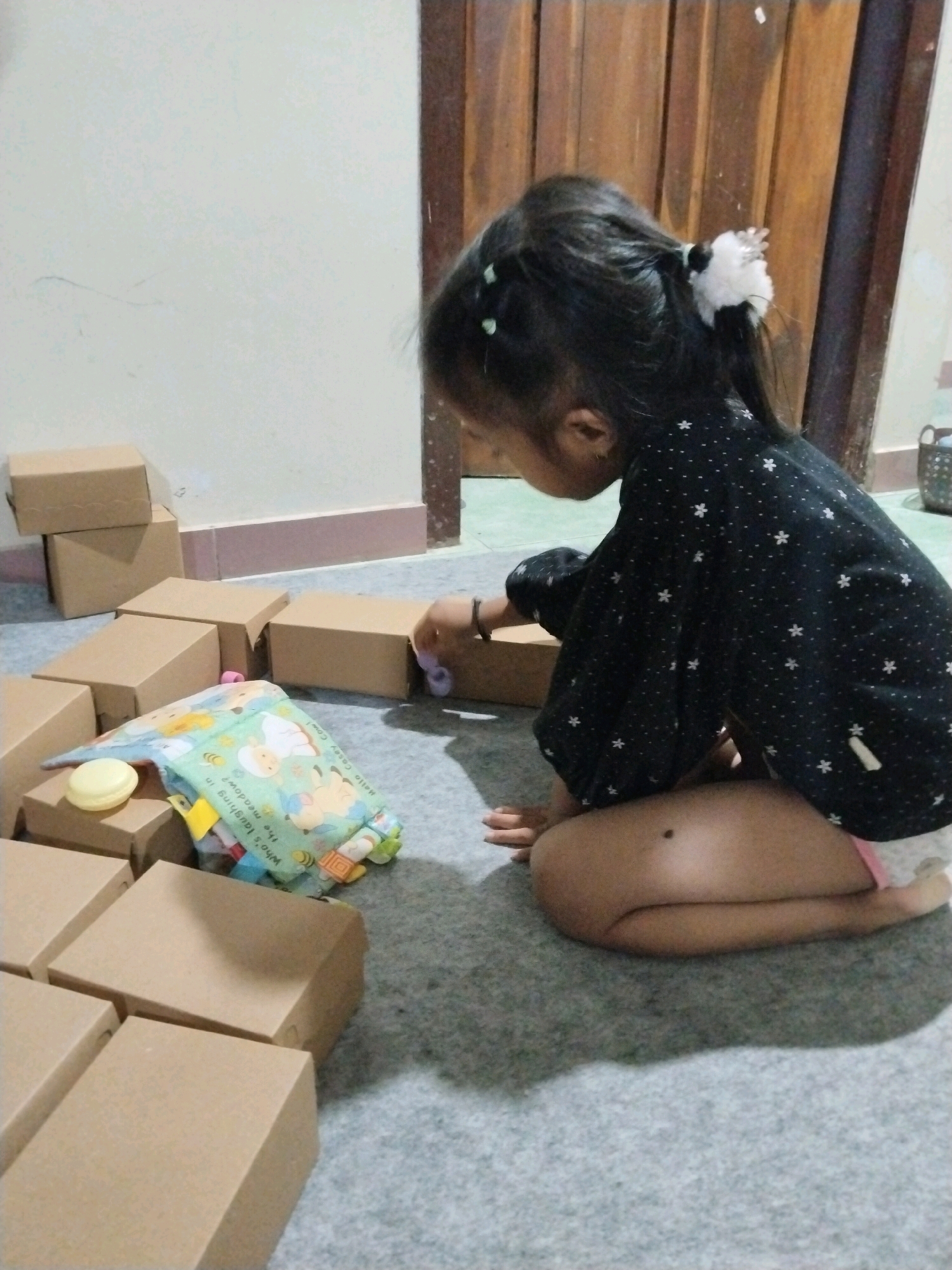 In Tasikmadu, Indonesia, a young girl is playing on the floor with cardboard boxes. She is wearing a black shirt with white stars and white socks. The girl is sitting on her knees, looking at the boxes, and her hands are near the boxes. There are several cardboard boxes on the floor, arranged in a scattered manner. A colorful soft book is also on the floor near the boxes. The room has a plain wall and a wooden door in the background. The lighting is dim, suggesting it is likely indoors.