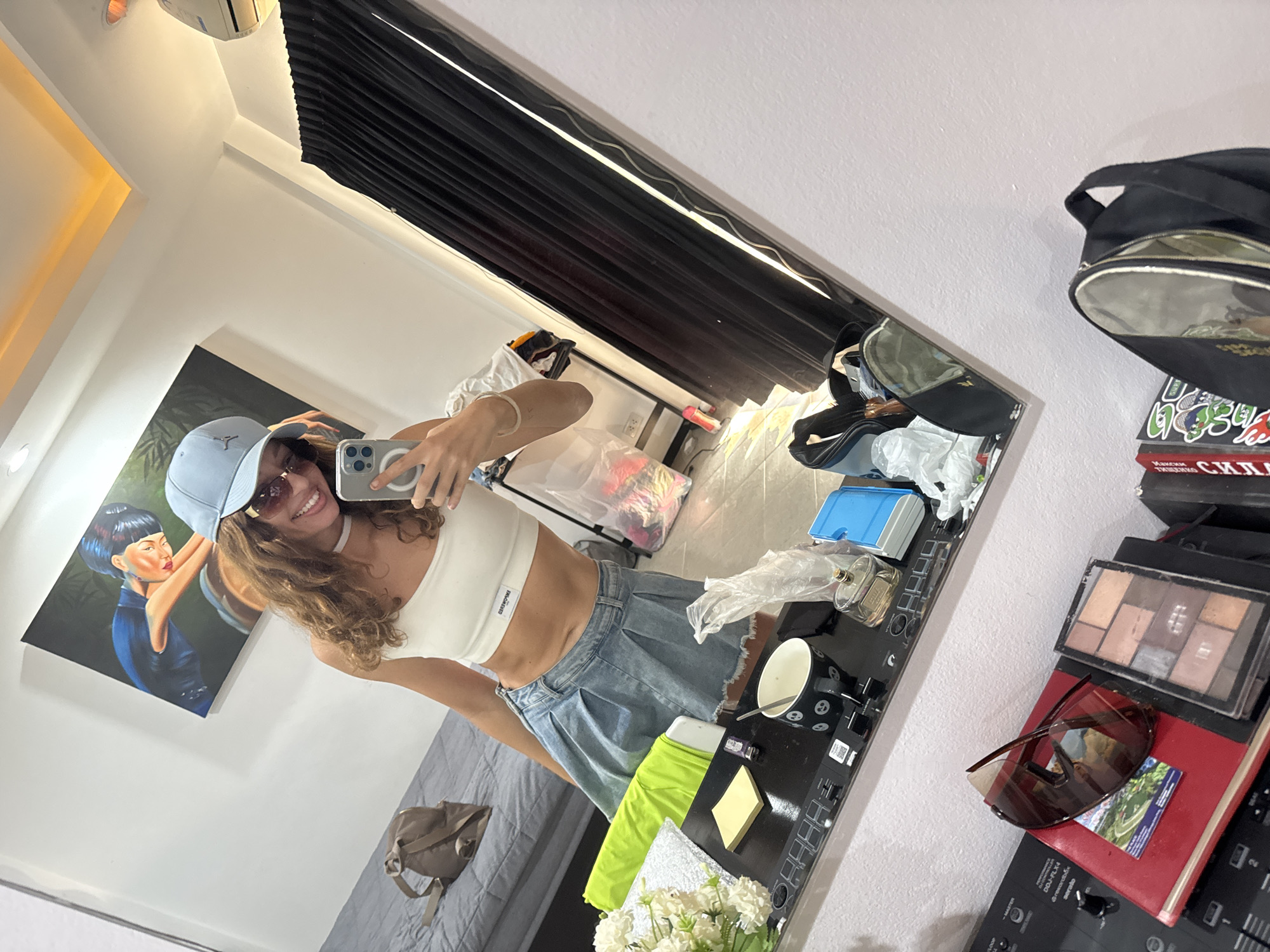 A woman is taking a selfie in a mirror. The woman is wearing a white top, light blue denim shorts, a light blue baseball cap, and sunglasses. The reflection shows a painting on the wall, a grey sofa, and personal belongings on a surface beside the mirror including makeup, sunglasses, and a bag. The location is potentially Baan Chaweng, Thailand based on general characteristics of the setting.