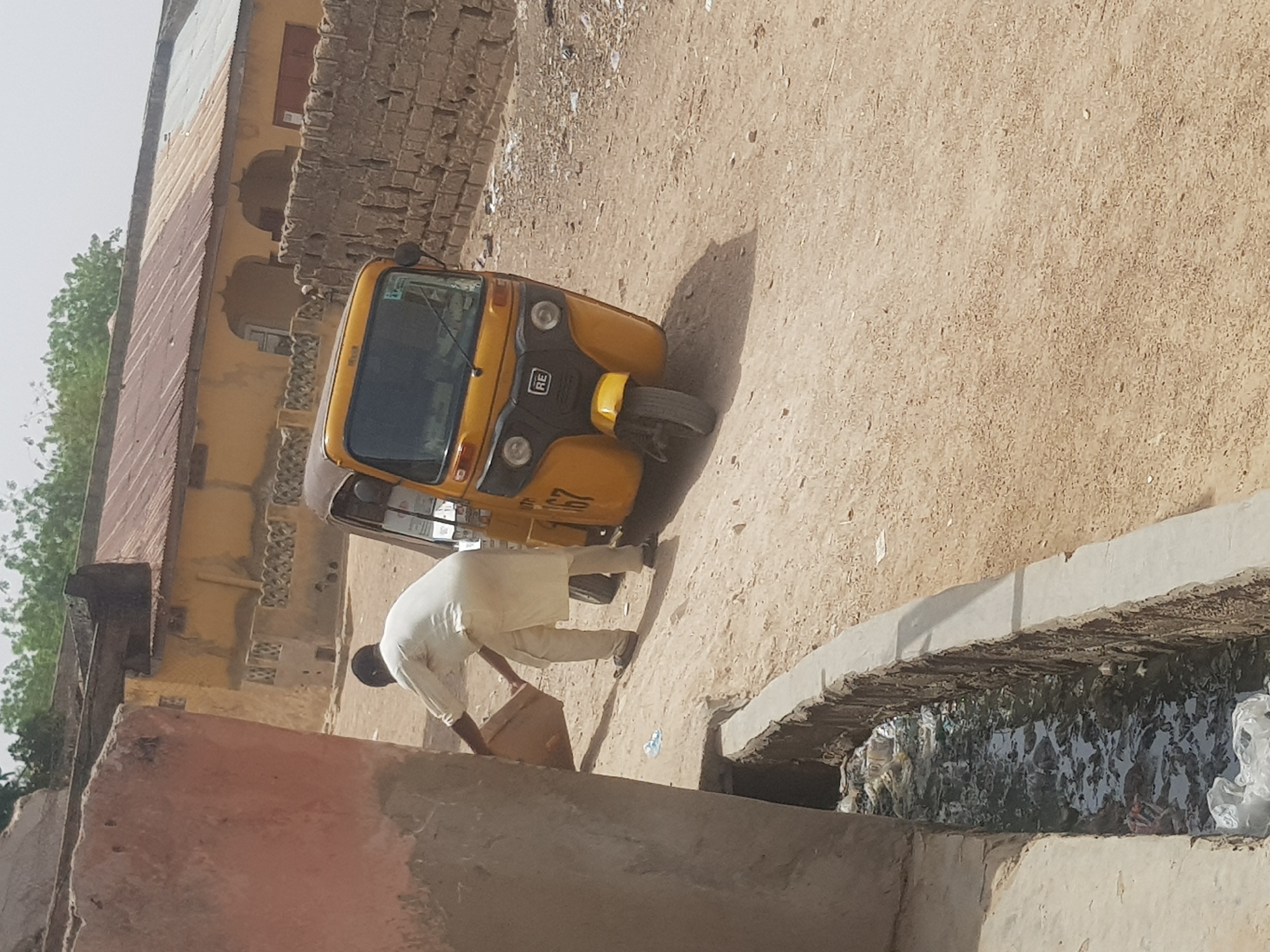 A man in a white tunic is dumping cardboard into a canal alongside a road. A yellow tricycle rickshaw is nearby. The canal is filled with stagnant water and rubbish. The road is unpaved. Buildings are visible in the background. The location is Dutsinma B, Nigeria.