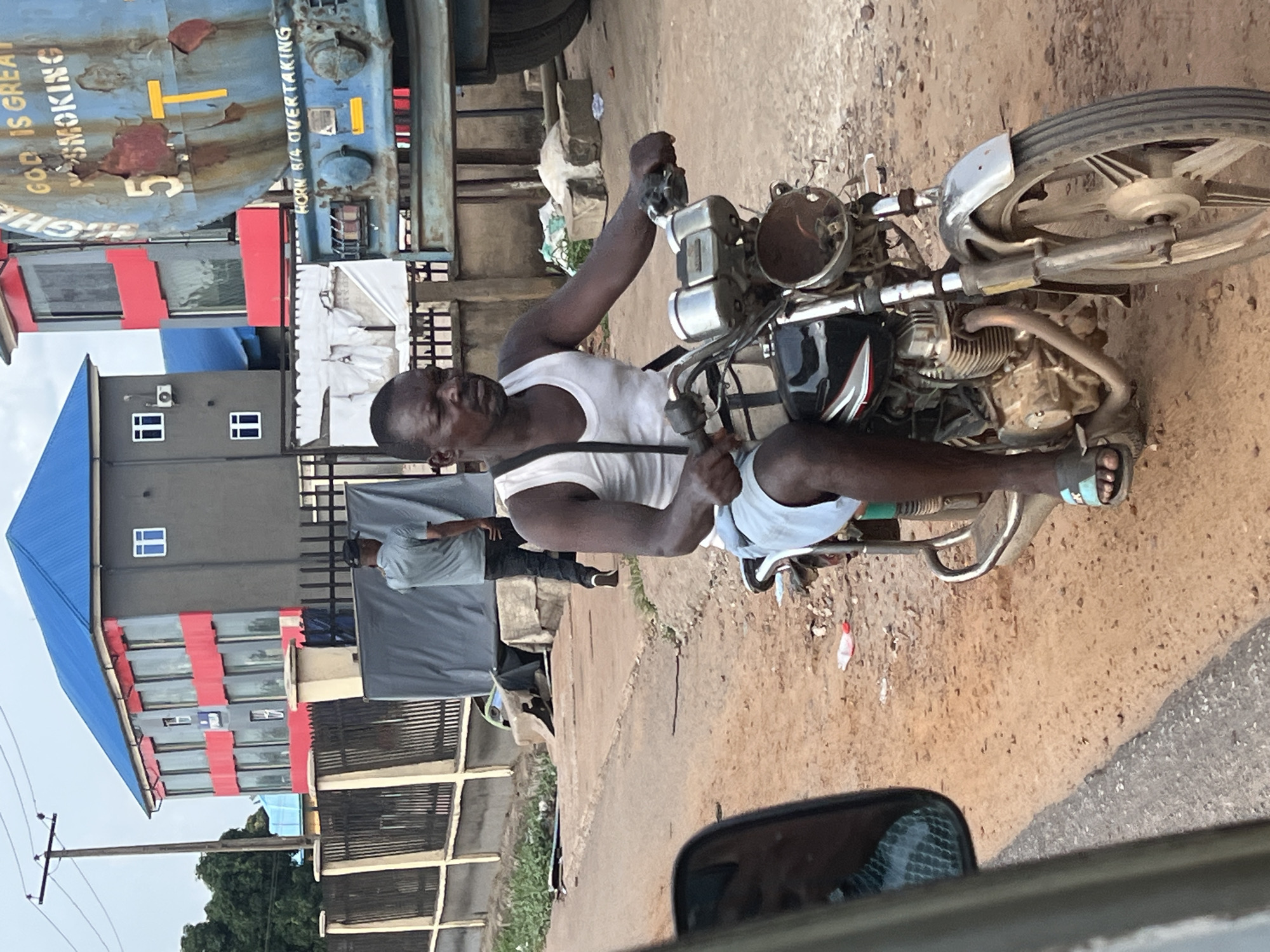 The image shows a man sitting on a motorcycle on a dirt road. He is wearing a white tank top and light blue shorts. The motorcycle is old and rusty, with a black and silver color scheme. Behind the man, there is a building with a blue roof and red stripes on the walls. To the left of the motorcycle, there is a truck. The photo was taken from inside a car, as a side mirror is visible in the bottom right corner. The location is an unidentified city in an unknown region.