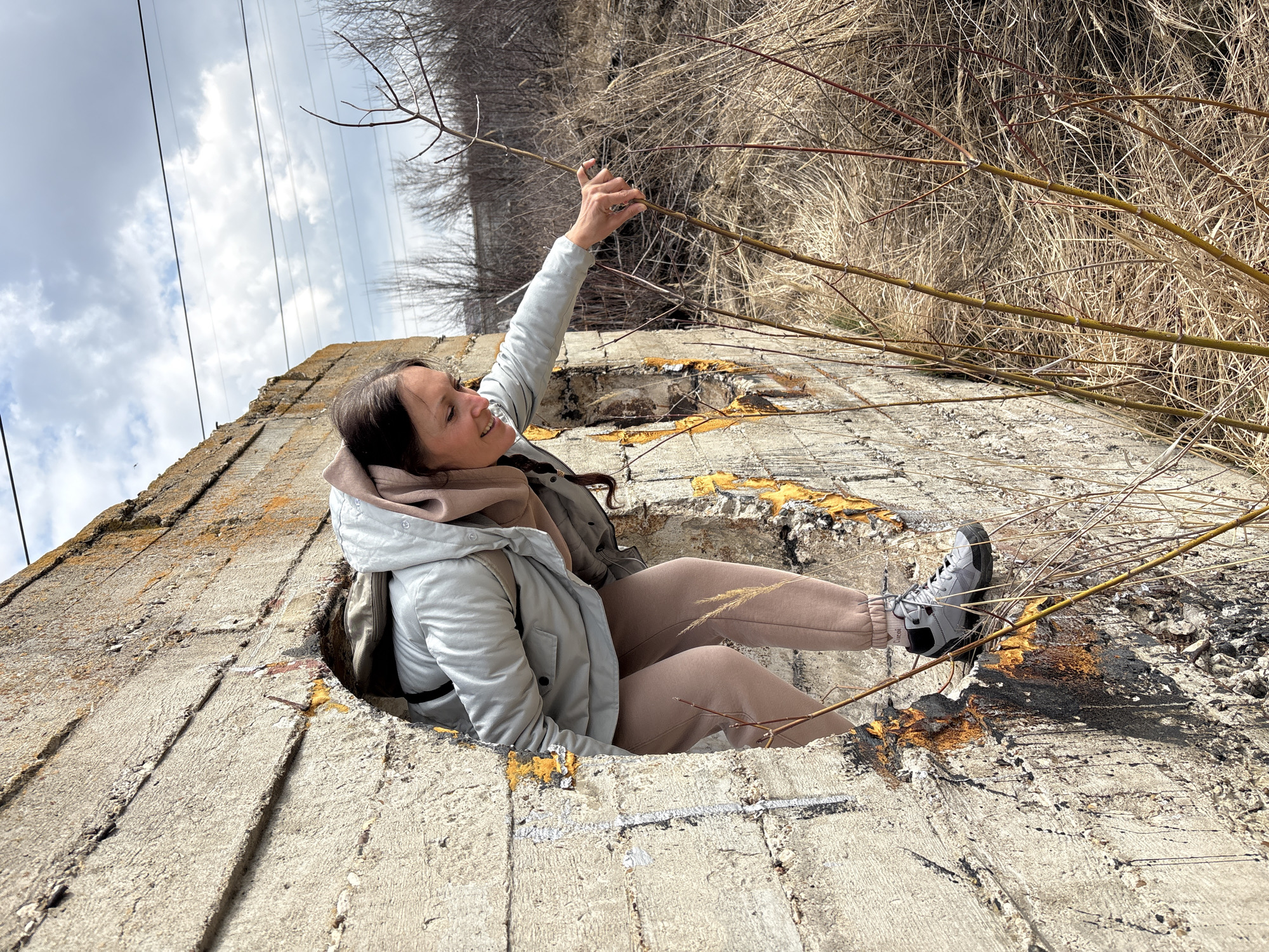 A woman is sitting in a hole in a concrete structure outdoors. The concrete structure is light grey, with areas of orange and yellow staining. The woman has dark hair and is wearing a light grey jacket, a beige hoodie, and beige sweatpants. She is wearing grey and black sneakers. Her right arm is raised, and her hand is holding a branch. There are many bare branches around her. The background is a mix of dry grass and leafless trees. Overhead, power lines are visible against a cloudy sky. The location is Ulyanovsk, Russia.