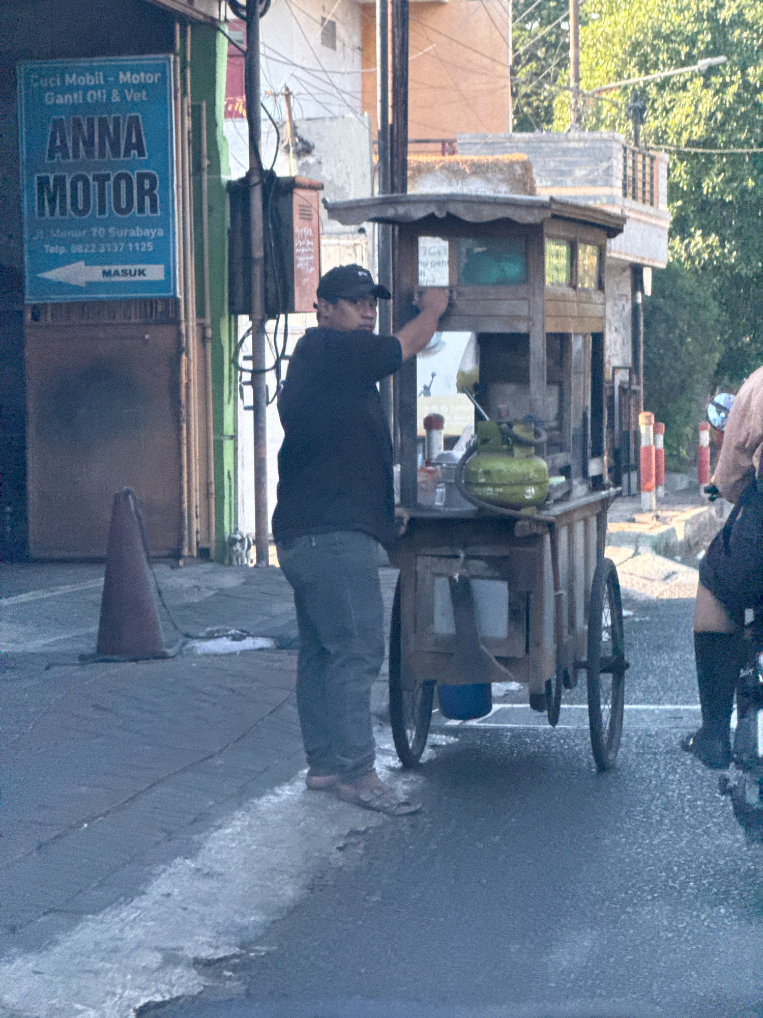 The image depicts a street scene in Surabaya, Indonesia, with a man operating a food cart. The sign behind him, "ANNA MOTOR," indicates a business that services cars and motorcycles. The man, wearing a dark t-shirt, cap, and jeans, appears to be preparing or serving food from the cart, which is equipped with a green gas cylinder and various containers. The cart has wooden construction and two wheels visible. In the background, there are buildings and trees. A traffic cone is visible on the left side of the image. Another person, partially visible on the right, is on a motorcycle. The lighting suggests it is daytime, possibly late afternoon or early morning, given the shadows. The environment appears to be an urban street with a paved surface.