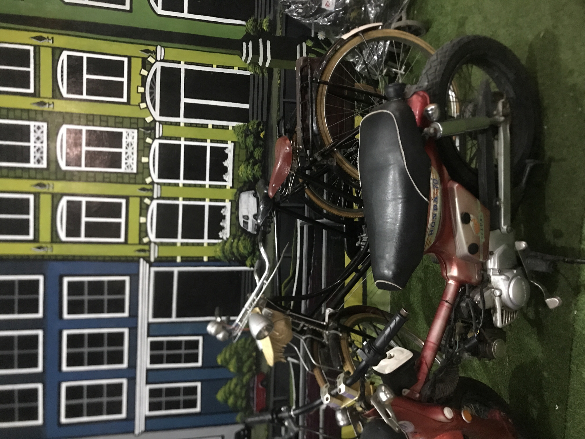 A motorcycle and bicycle are parked in front of a building facade. The motorcycle is red and silver with a black seat. The bicycle is black with brown tires. The building facade is painted with different colors and has windows. The ground is covered with green artificial grass. The location is Keudeteupinpunti, Indonesia.