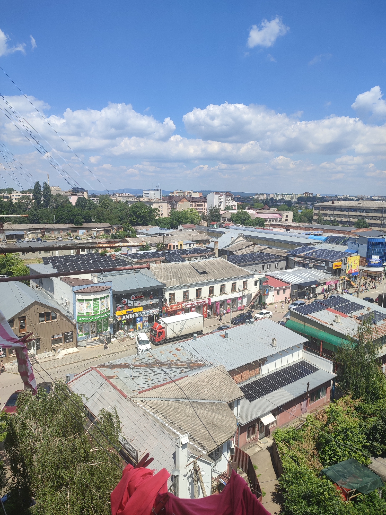 An elevated, eye-level, full shot captures a view over a densely built area of Chernivtsi, Ukraine, on a sunny day with scattered white clouds against a bright blue sky. The foreground features the tops of trees and laundry hanging on a line, obscuring a small portion of the image. The mid-ground is dominated by a collection of buildings with various roof types, including metal, shingled, and some covered with solar panels. Numerous commercial establishments are visible, with signs in Cyrillic script, such as "АНТЕКА ОПТОВИЙ" and "ANDI-GROUP." A red truck, a white van, and several cars are parked on a street running through the scene, indicating it is a commercial district. In the background, a cityscape unfolds with more buildings of varying heights and architectural styles, interspersed with trees, and a distant view of hills. The overall impression is that of a lived-in urban environment with a mix of older and newer infrastructure, including the prominent use of solar energy.
