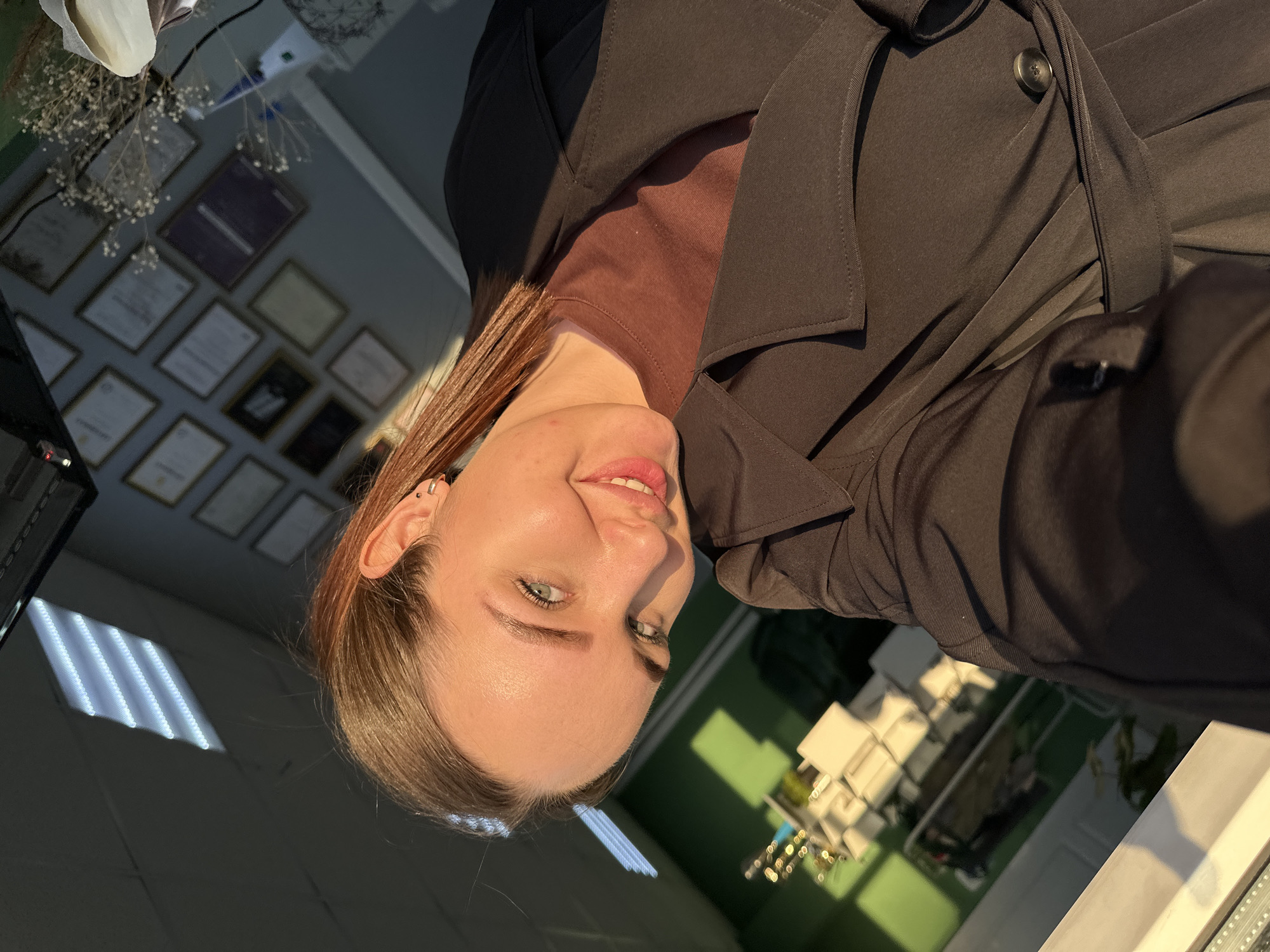 A woman is taking a selfie indoors. She has fair skin, light-colored eyes, and light brown hair. She is wearing a brown turtleneck shirt and a dark-colored trench coat. The background shows a wall with numerous framed certificates or diplomas. There is also a light source in the upper left, potentially fluorescent lighting. In the lower right, a building or structure is visible. The photo appears to have been taken at a slight angle and the location cannot be determined to be the city of Chaikovsky, Russia based on the details available in the picture.