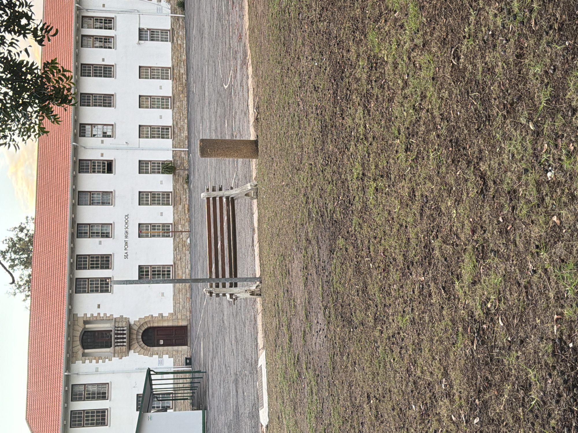 The image displays the exterior of a building identified as "SEA POINT HIGH SCHOOL," located in Cape Town, South Africa. The scene is captured during the daytime under natural light, with a bright sky visible.
The primary subject is a multi-story white building with a distinctive red tiled roof. The building features numerous rectangular windows with light-colored frames, arranged in rows. Its architecture includes decorative stone detailing, particularly around a prominent entrance on the lower floor, which has an arched wooden door and a small balcony above it. The name "SEA POINT HIGH SCHOOL" is written vertically on the building's facade on the right side of the main entrance.
In front of the building, there is a paved area, possibly a courtyard or pathway, made of what appears to be asphalt or dark concrete. Adjacent to this paved area, to the right, is a large patch of grass. The grass appears somewhat sparse or dry, with visible soil and scattered dry foliage.
Within the grassy area, a single large, cylindrical brown bollard or pillar stands. To its left, partially visible, is a wooden slatted bench with a light-colored frame. On the far left of the image, green tree branches with leaves extend into the frame, suggesting the presence of trees nearby. No people or specific activities are visible in the captured moment, presenting a quiet, unpopulated view of the school grounds.