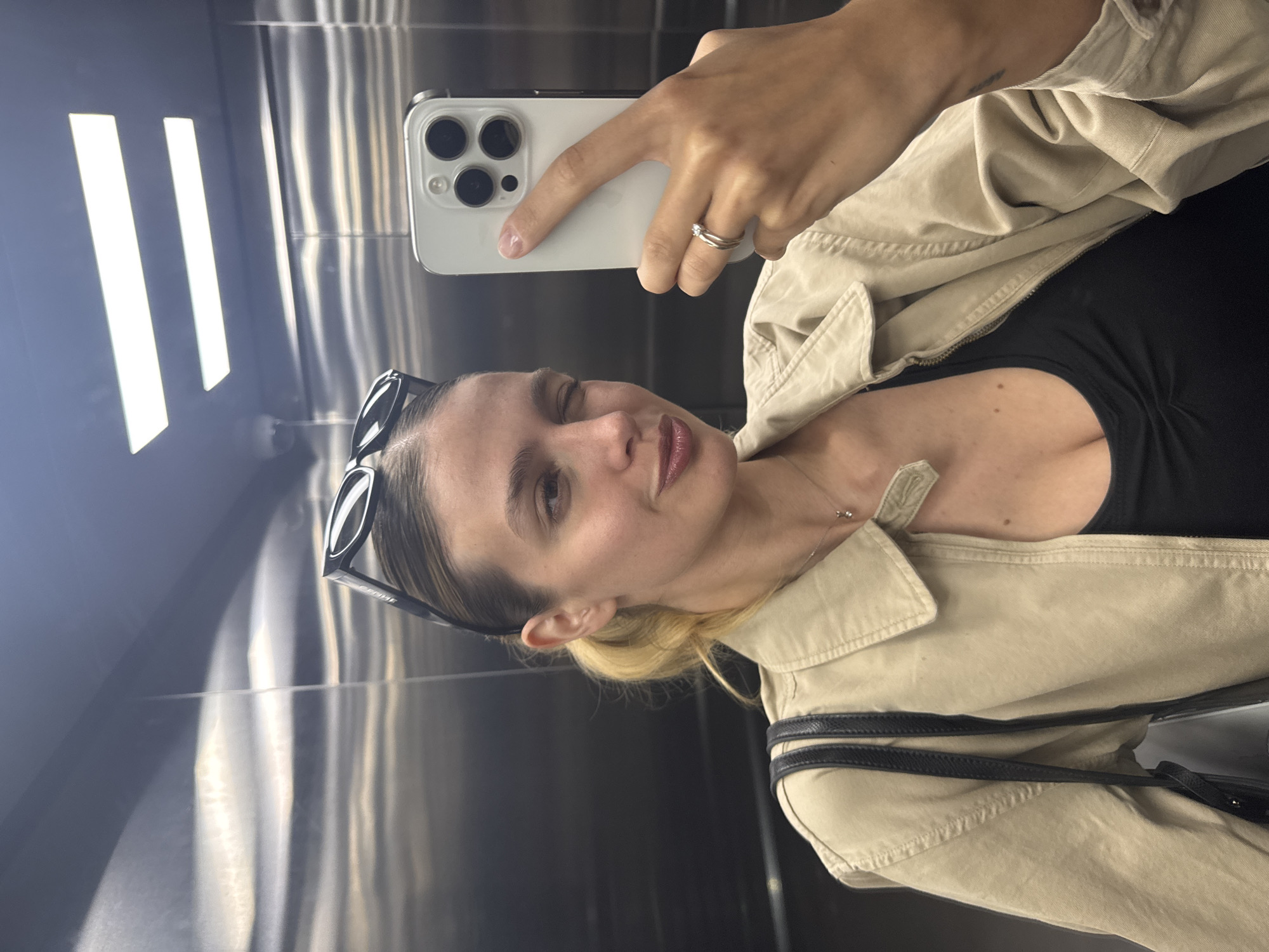 A woman takes a selfie in a reflective elevator. She is holding a smartphone with multiple camera lenses in her right hand. She is wearing a beige jacket over a black top. Sunglasses are perched on top of her head. The elevator has a metallic, reflective interior with vertical lights. The location is unknown.
