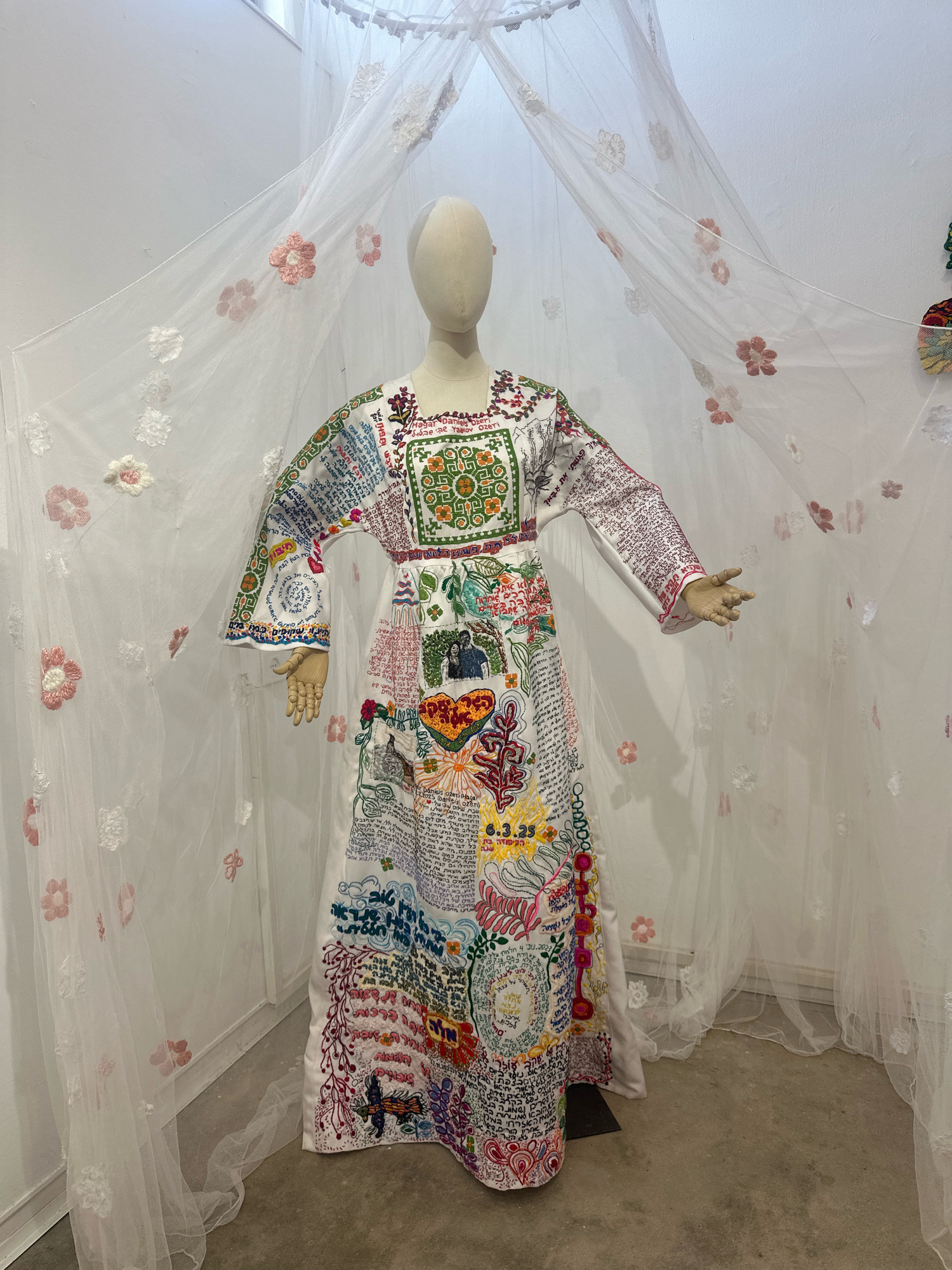 A mannequin is displayed wearing an elaborately embroidered white dress adorned with various colorful patterns, text, and imagery. The dress features a high neckline, long sleeves, and a floor-length skirt, all covered in intricate needlework. The mannequin stands centrally within a space that appears to be an art installation or exhibition. Behind the mannequin, a sheer white canopy or curtain, decorated with embroidered flowers in pink and white, drapes from above, creating a soft, ethereal backdrop. The floor is concrete, and the walls are painted white. The overall impression is one of artistic presentation, likely in a gallery or museum setting within the city of Mate Asher Regional Council, Israel.