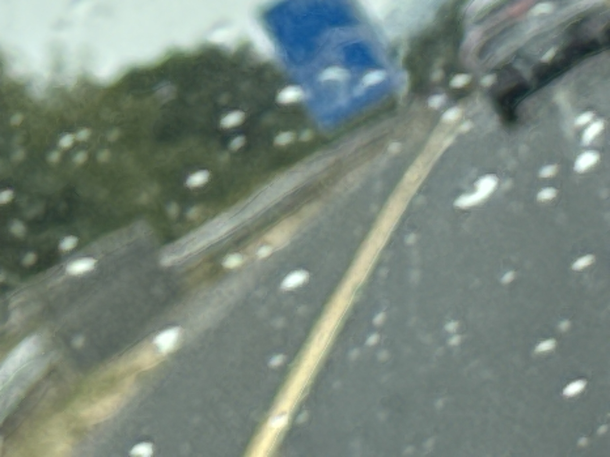 The image depicts a blurry scene, possibly taken through a wet window or windshield, showing elements of an outdoor environment. In the upper portion of the image, a blurred green area suggests vegetation or trees. A blue object, potentially a vehicle or a structure, is visible in the middle-left, partially obscured by the blur. The foreground is dominated by a grey surface, likely asphalt, which appears to be a road or parking area, with a bright yellow line running diagonally across it, indicating a lane divider or road marking. Numerous small white spots are scattered throughout the image, which could be water droplets or reflections. The overall impression is of an overcast day, with low visibility and a sense of movement or observation from a stationary point. The context of Bennettsbridge, Ireland, suggests this scene is likely within or near the village, possibly on a road or near a public area.