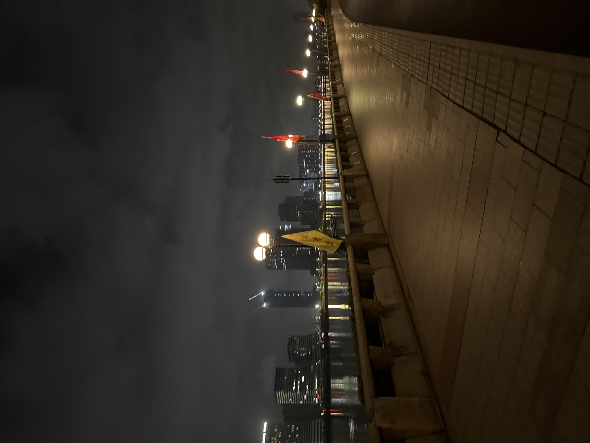 This is a night-time, vertical, eye-level shot of a waterfront promenade in Haizhu District, China. The promenade is paved with large, rectangular light-brown tiles, bordered by a textured strip of smaller, darker tiles. A low, stone wall with square indentations runs along the left side of the promenade, separating it from a body of water. Beyond the water, the city skyline is visible, illuminated by numerous lights. Several modern high-rise buildings, some with visible construction cranes, are prominent in the background. A series of streetlights with double globes line the promenade, casting a warm glow on the path. Red flags on poles are spaced along the walkway, and a yellow flag is attached to a lamppost in the foreground. The sky is dark and cloudy. The scene is devoid of people.