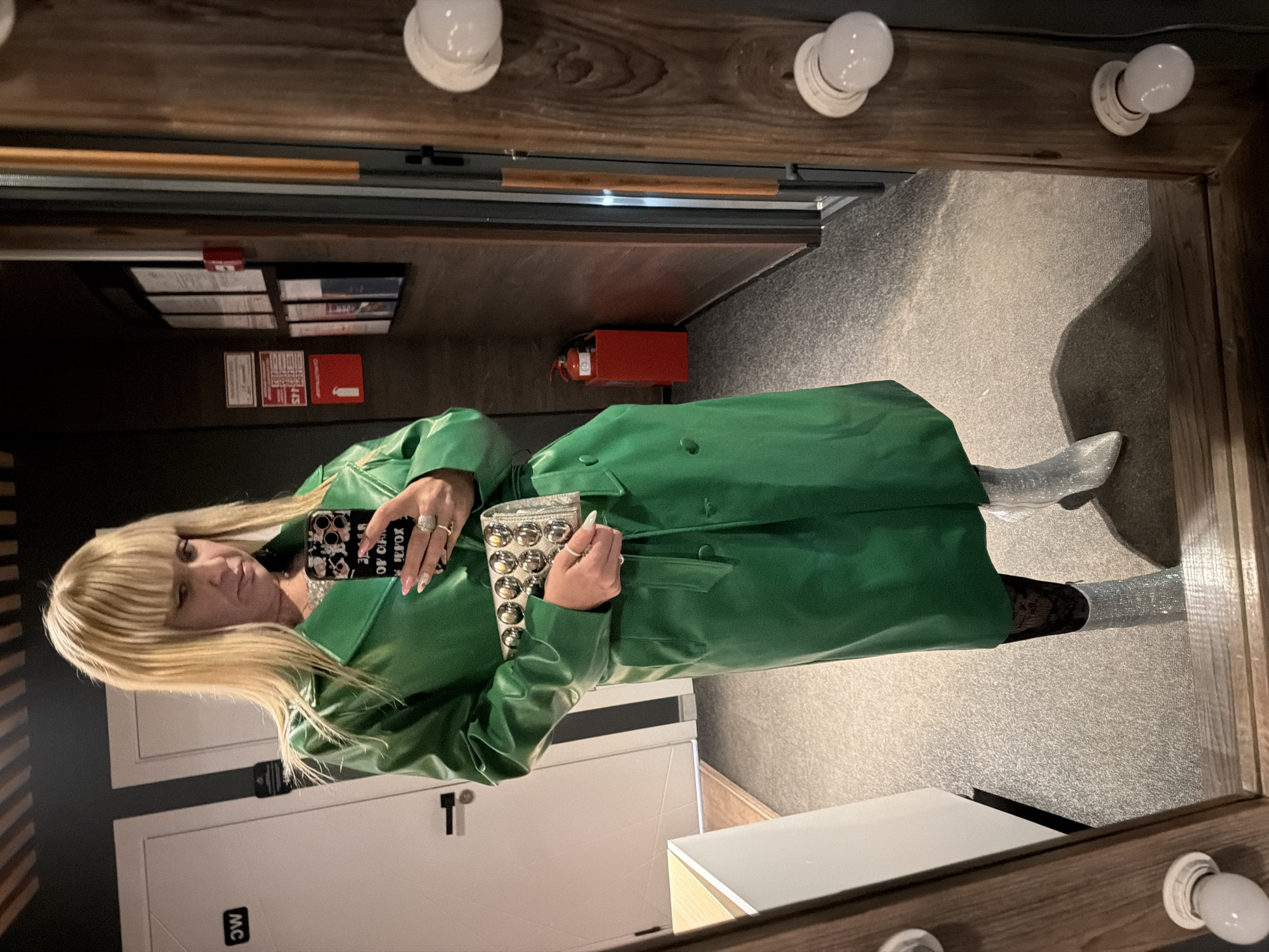 A person with blonde hair and a green trench coat is standing in a room with a large mirror. The room appears to be a dressing room or bathroom, judging by the vanity lights around the mirror. The person is holding a mobile phone and a studded clutch bag. The background includes a door with "MC" written on it, and what seems to be fire safety information posted on the wall. A fire extinguisher is visible as well. The exact location is difficult to determine without additional information, but the query requested the city of Chaikovsky, Russia.