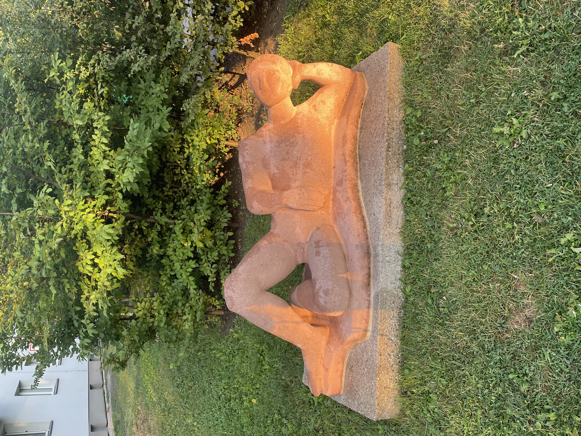 A terracotta-colored sculpture of a reclining nude figure rests on a concrete base in a grassy area. The figure is positioned on its left side with its legs bent and crossed. Its right arm is bent, with the hand supporting the head, which is tilted back. The sculpture is illuminated by warm, late afternoon sunlight, casting long shadows. Behind the sculpture, a dense green hedge provides a backdrop, and to the left, a portion of a light-colored building with white-framed windows is visible. The location is the garden of the Otto Wagner Church of St. Leopold in Vienna, Austria, specifically near the entrance.