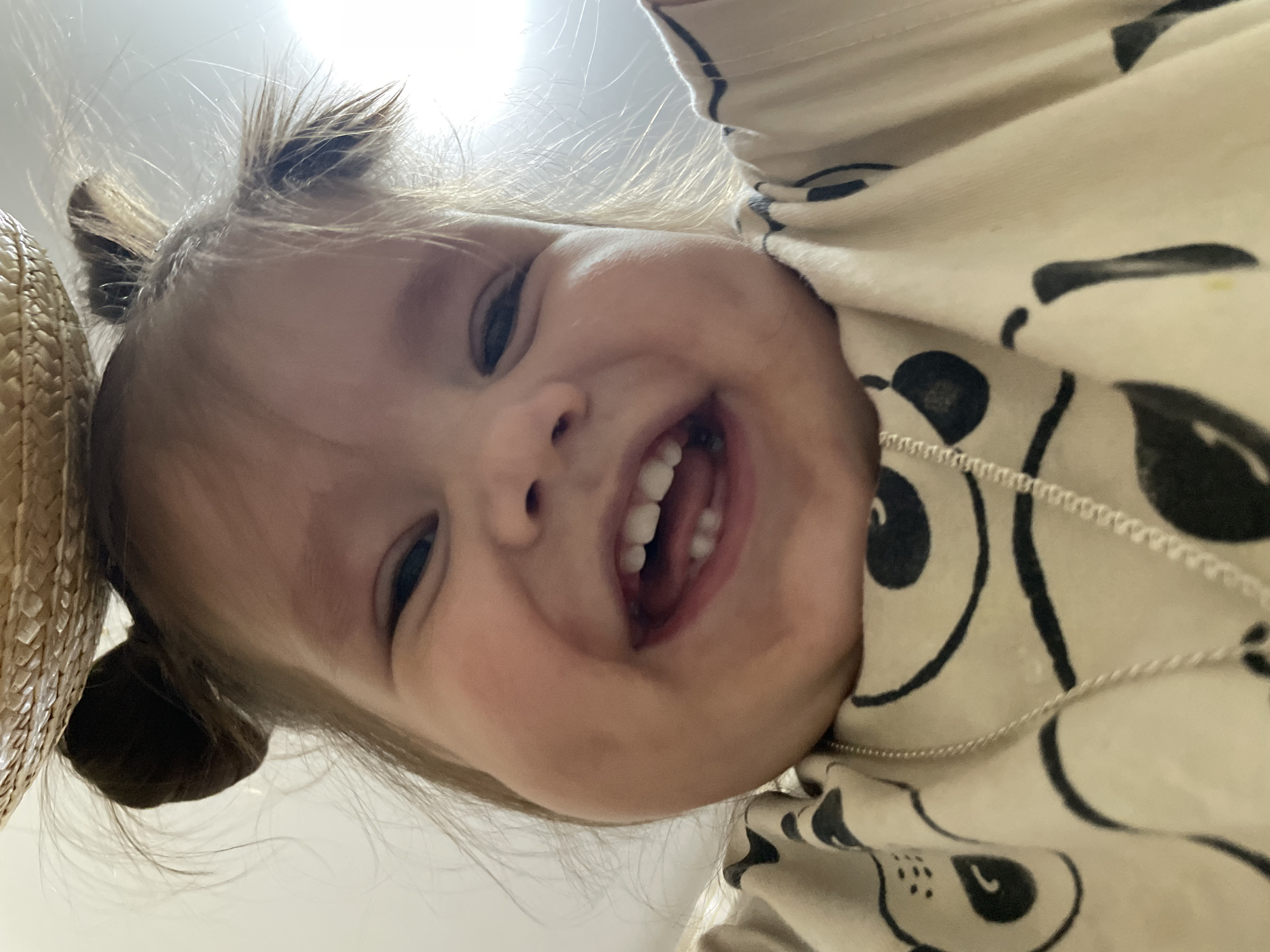 A young child with light skin and brown hair styled in two small buns is smiling directly at the camera. The child has visible teeth and a slightly upturned nose. A portion of a straw hat is visible on the left side of the frame. The child is wearing a light-colored garment with a black and white pattern. A thin silver necklace is also visible. The background is a plain, bright white. The location is not discernable from the image.