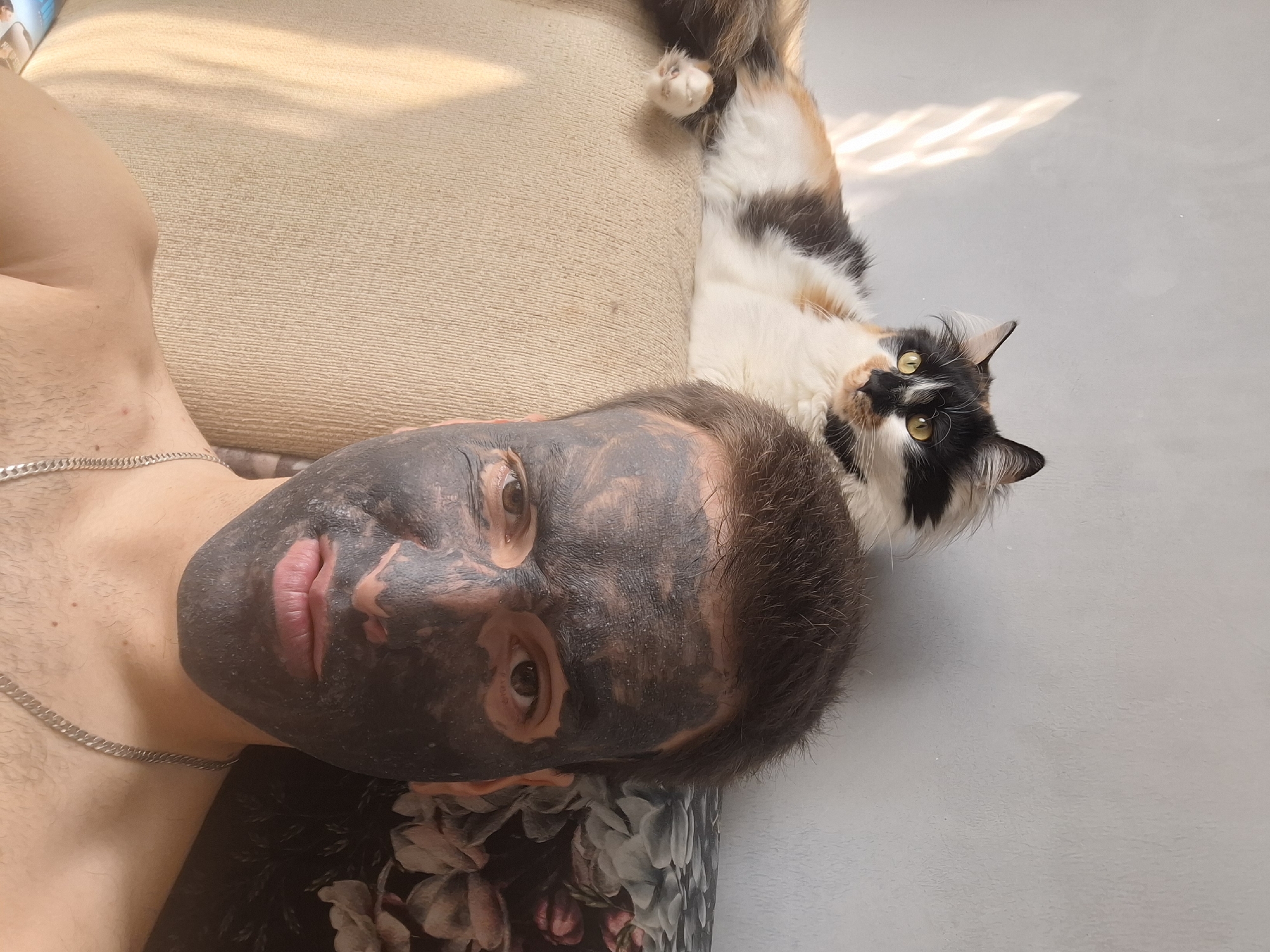 A man with a dark face mask lies on a patterned cushion next to a beige sofa. A calico cat is lying on a gray surface. The location appears to be indoors. The city of Sofia, Bulgaria cannot be confirmed from the information provided.