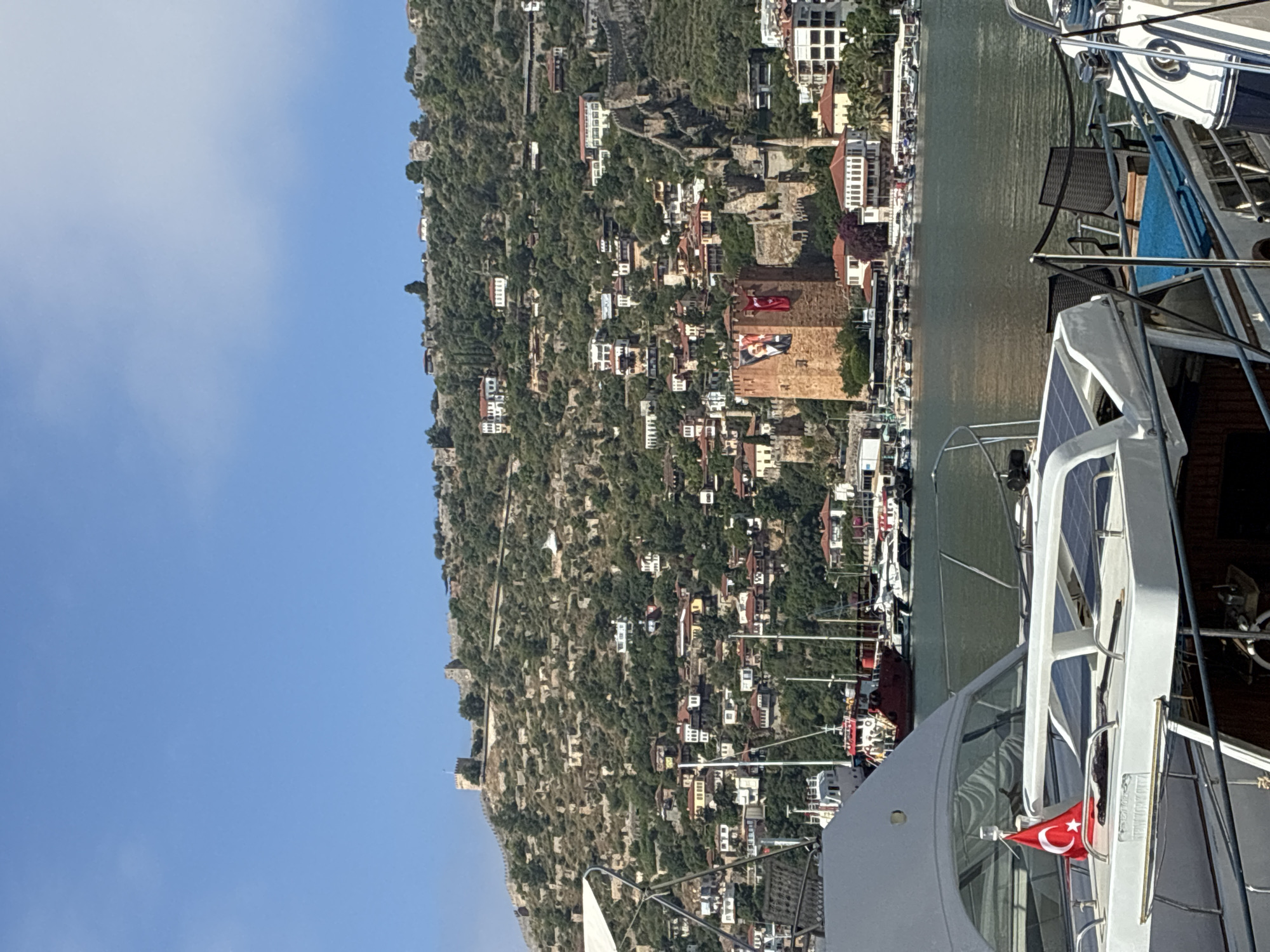 The image depicts a sunny day in the city of Alanya, Turkey, showcasing a scenic view of the Alanya Castle complex overlooking the harbor. In the foreground, a white yacht is docked, featuring a small Turkish flag planted on its deck and solar panels visible on its roof. The yacht's interior is partially visible, revealing seating and other boat amenities. Beyond the yacht, the Alanya harbor is filled with various boats, including a red ferry and several smaller vessels.
Dominating the background is the historic Alanya Castle, perched atop a steep, verdant hill. The castle is characterized by its imposing stone walls and towers, with scattered ruins and buildings visible along the hillside. A prominent red tower, part of the castle complex, flies a large Turkish flag and a portrait, possibly of Mustafa Kemal Atatürk. Numerous houses with red-tiled roofs are nestled into the hillside, interspersed with dense green vegetation. The sky is a clear, bright blue with a few wispy clouds. The water of the harbor is calm and reflects the surrounding scenery.