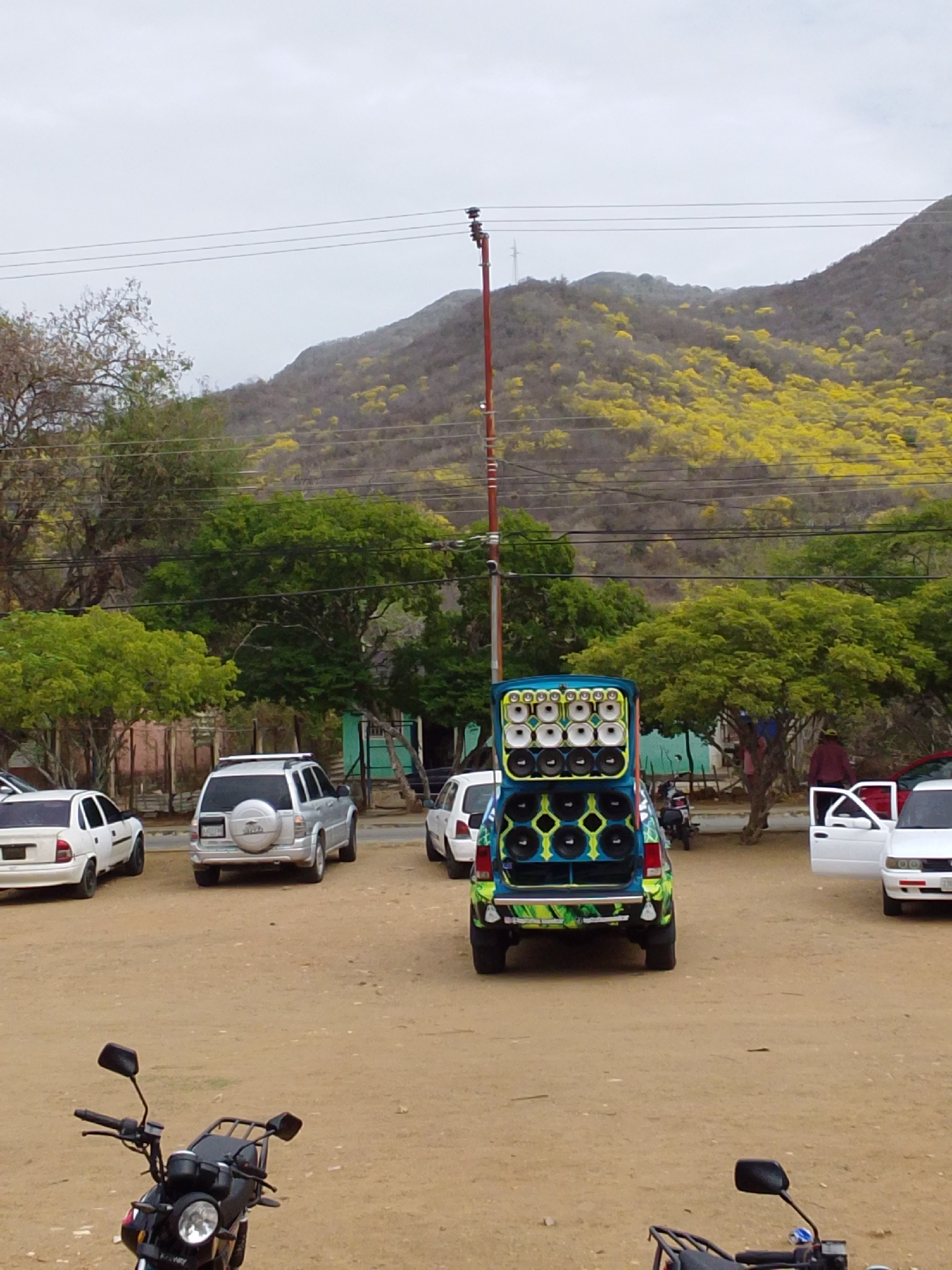 The image depicts an outdoor scene in an unpaved, dirt area under an overcast sky. In the foreground, the handlebars and front headlight of a black motorcycle are partially visible on the bottom left, with another motorcycle partially visible on the bottom right.
The central subject is the rear of a custom-modified light blue and green van or small SUV, parked facing away from the viewer. Its rear hatch is open, revealing a large array of speakers. The top section contains eight smaller circular speakers arranged in two rows of four. The bottom section features six larger circular speakers arranged in two rows of three, set within a frame adorned with bright green and yellow geometric patterns. The lower rear bumper of the vehicle also exhibits a similar green and yellow design.
To the left of the speaker-equipped vehicle, a white sedan is parked, facing left. Further behind and to the left, a silver SUV with a rear-mounted spare tire is parked, facing right.
To the right of the speaker vehicle, a white two-door car is parked with both its driver-side and passenger-side doors open. A person wearing a red shirt and a hat is visible standing next to the driver's side door of this white car. Another person is partially visible behind the speaker vehicle, near a tree.
The background features a range of hills or mountains covered with vegetation. A significant portion of these trees are blooming with bright yellow flowers, creating a vibrant pattern against the muted green and brown foliage. Several green-leafed trees are scattered across the mid-ground. A red utility pole with multiple cross arms and numerous overhead power or communication lines extends vertically from the mid-ground into the upper portion of the image. A light blue building or structure is partially visible behind the trees in the mid-ground.
The ground is predominantly light brown, unpaved soil, suggesting a rural or semi-urban setting. The location is in the city of Unknown, Unknown.