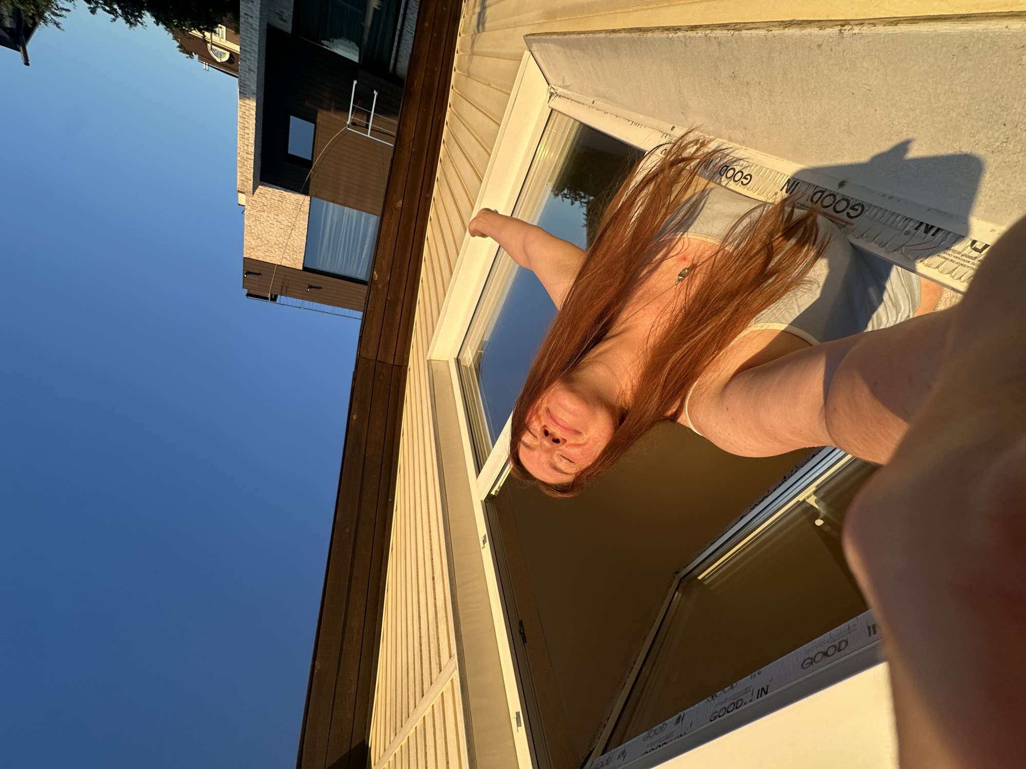 A young woman with long auburn hair is leaning out of a window, her face tilted up towards the camera and her eyes closed. She has a bright, happy expression and appears to be enjoying the moment. The sun is shining brightly, casting a warm glow on her face and hair. Her hair is blowing in the wind, adding to the sense of freedom and joy.
She is wearing a light-colored top and what appears to be shorts. There is a necklace with a small pendant around her neck. The window frame is white, and there are some labels with the text "GOOD. IN" visible on the bottom edge of the window.
The scene is set against a backdrop of a modern building with a light-colored, vertical siding. Adjacent to this building, there is a darker building with a wooden exterior. The sky is a clear, vibrant blue, suggesting a beautiful, sunny day. The sunlight indicates it is likely late afternoon or early evening. The overall mood is cheerful and relaxed, capturing a moment of simple pleasure.