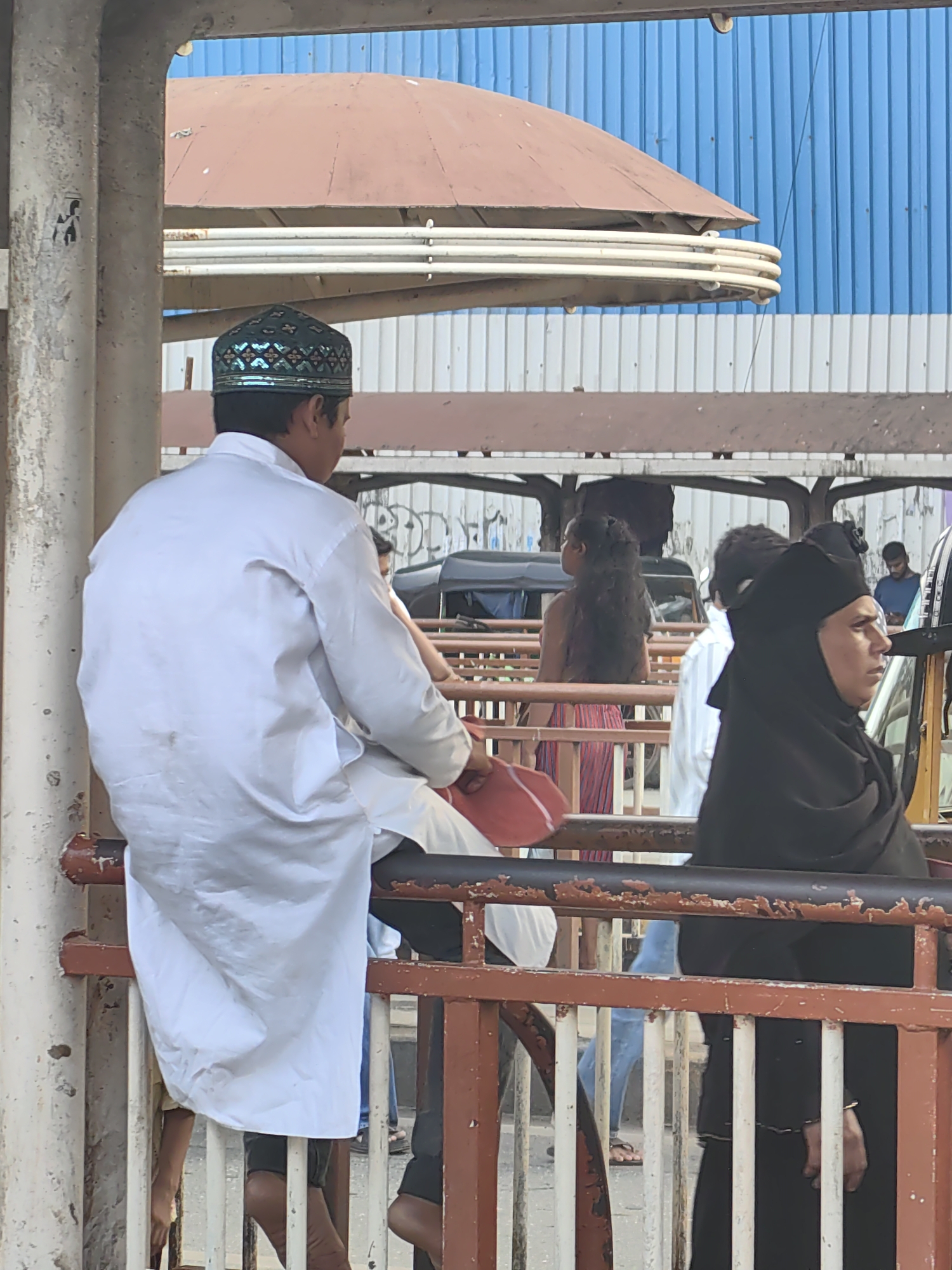 The image captures a street scene in Mumbai, India, likely near a public transport hub given the presence of railings, a bus shelter structure, and what appears to be a queue or waiting area. In the foreground, a man wearing a white kurta and a decorative embroidered cap is seated, facing away from the camera. To his right, a woman in a black hijab is visible, looking to the right. Behind them, several other individuals are present, including a woman with long, dark, curly hair wearing a striped skirt. Vehicles, possibly auto-rickshaws or small buses, are discernible in the background, suggesting an active urban environment. The overall setting appears to be outdoors, with a corrugated blue metal structure forming the backdrop. The lighting suggests it is daytime.