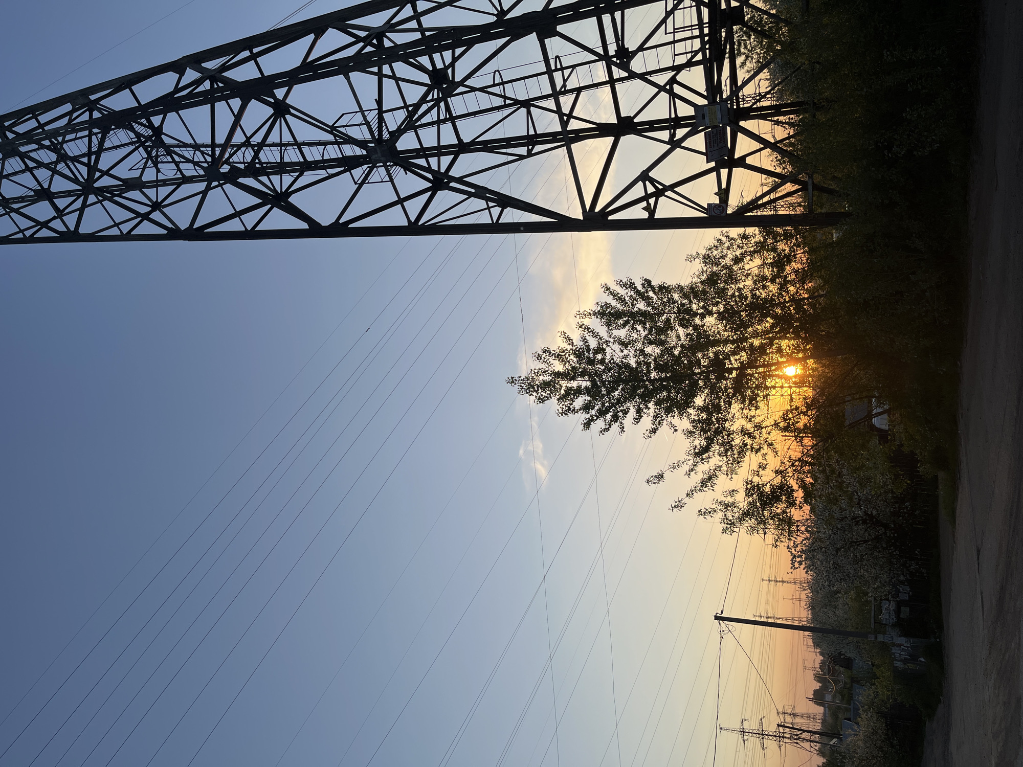 The image depicts a view in Chaikovsky, Russia, looking upwards towards an electricity pylon. The pylon's steel framework dominates the upper left of the frame. Power lines stretch across the sky. The setting sun is partially obscured by trees and vegetation on the right side of the image, creating a warm, orange glow. Other utility poles and infrastructure are visible in the distance, suggesting an urban or suburban setting. The ground is visible at the bottom of the frame, indicating a roadside location.