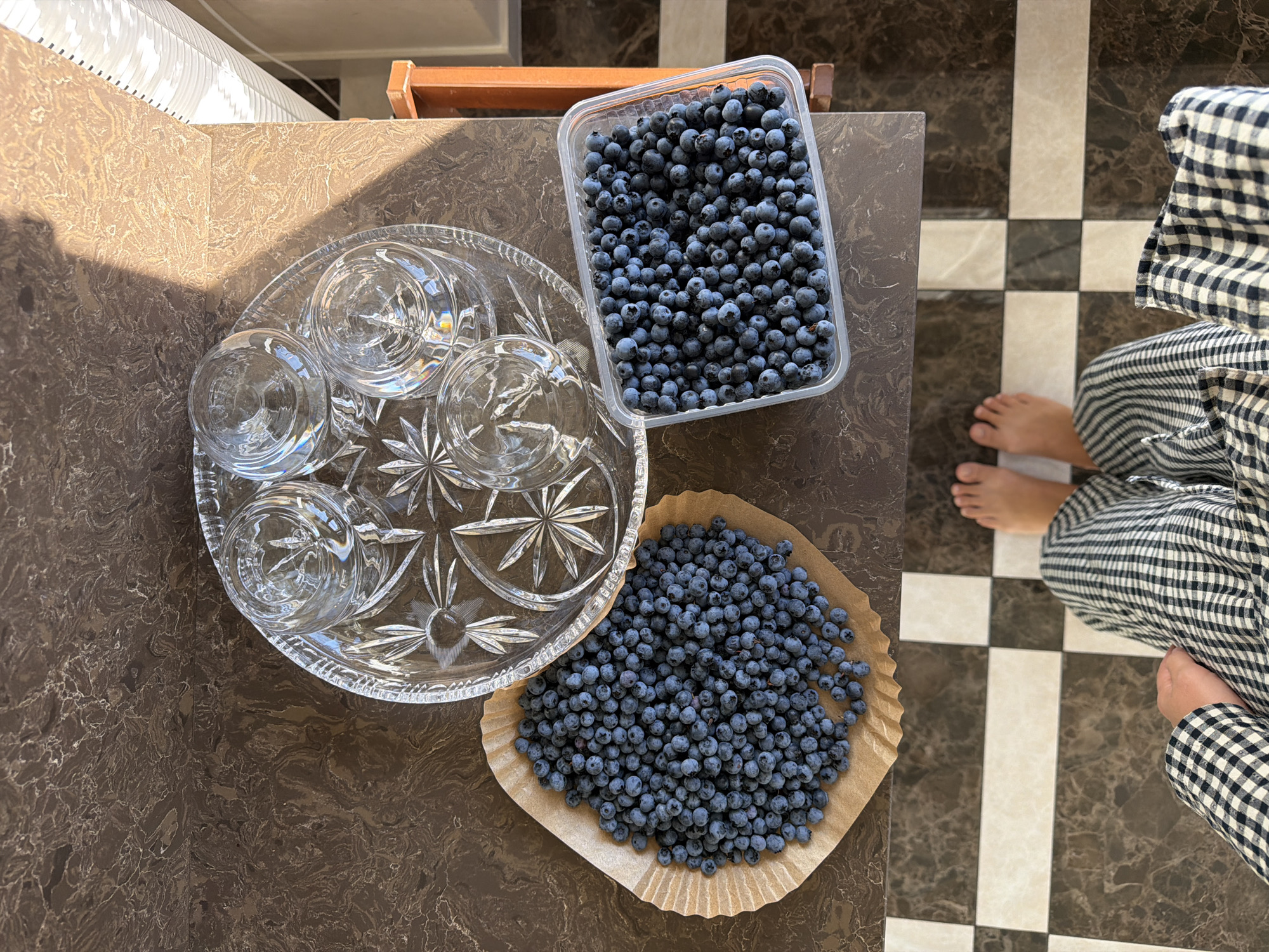A high-angle, close-up shot captures a variety of items arranged on a dark brown, marbled countertop, with a person's feet and legs clad in black and white checkered pants visible at the bottom of the frame. To the left, a large pile of fresh blueberries is spread out on a circular piece of brown parchment paper. To the right of the blueberries, a clear plastic container is filled with more blueberries. In the upper center, a crystal serving tray holds four clear drinking glasses, each partially filled with a pale blue liquid, suggesting they might contain a beverage. The countertop exhibits a swirling, marbled pattern with veins of lighter brown and white. Sunlight streams in from the right, casting a distinct shadow across the countertop and the serving tray, highlighting the texture of the surfaces. The floor below is tiled with a checkered pattern of dark brown and white squares. The overall impression is that of a domestic setting, likely a kitchen or dining area, possibly in Mishutkino, Russia, where fresh blueberries are being prepared for consumption.