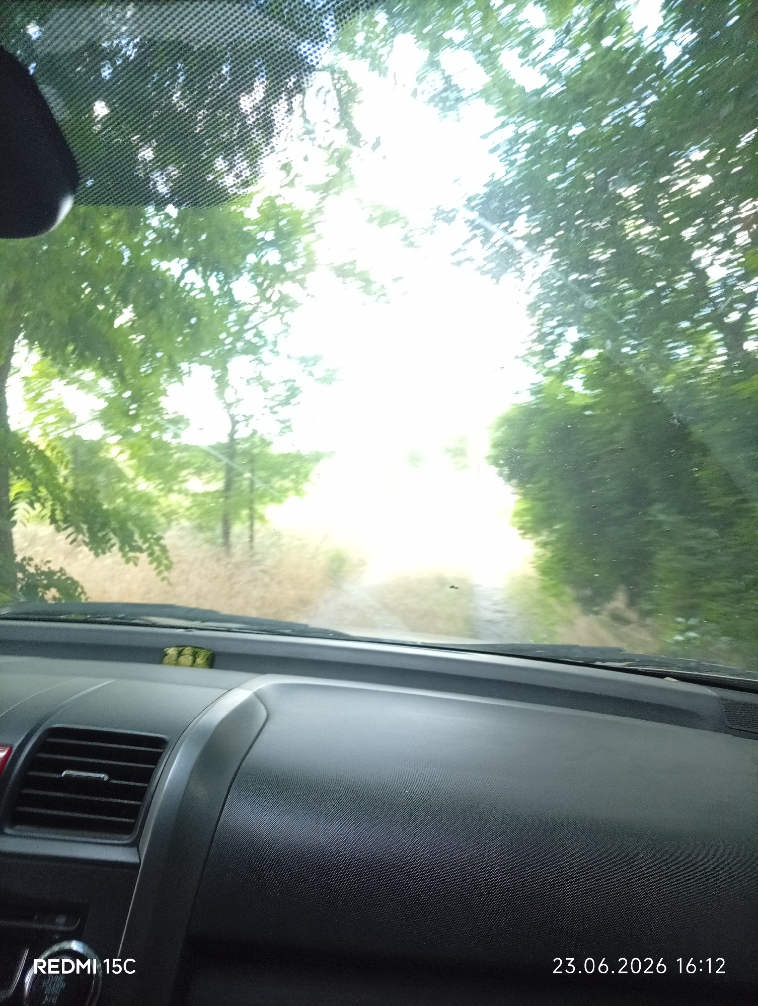 The image, taken from inside a vehicle, shows a dashboard and the view through the windshield. The windshield is partially obscured by the interior of the car, including a rearview mirror on the left and part of the dashboard with an air vent. Outside the car, the view is of a dirt road or path lined with trees and foliage, leading into a bright, sunlit area. The date and time stamp in the bottom right corner indicates "23.06.2026 16:12". The lower left corner shows the text "REDMI 15C", identifying the device used to capture the image. The scene appears to be a rural or natural setting, possibly a park or wooded area. There are no people visible in the image, and the focus is on the interior and exterior view of the vehicle. The location is identified as Mykolaivka, Ukraine.