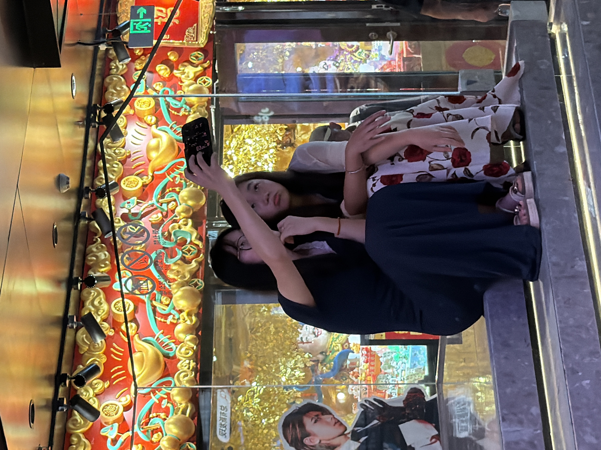 Two young Asian females are seated on stone steps in front of a building entrance. The female on the left, wearing glasses and a dark blue dress, is holding up a smartphone to take a selfie. The female on the right is wearing a white dress with a red floral print. Behind them is a glass-enclosed display with red and gold ornate decorations featuring Chinese motifs. Above the display, a green exit sign with an arrow points to the right. The scene appears to be outdoors in an urban environment, possibly in the Yuzhong District of China, indicated by the traditional Chinese decorations and signage. The lighting suggests it is evening.