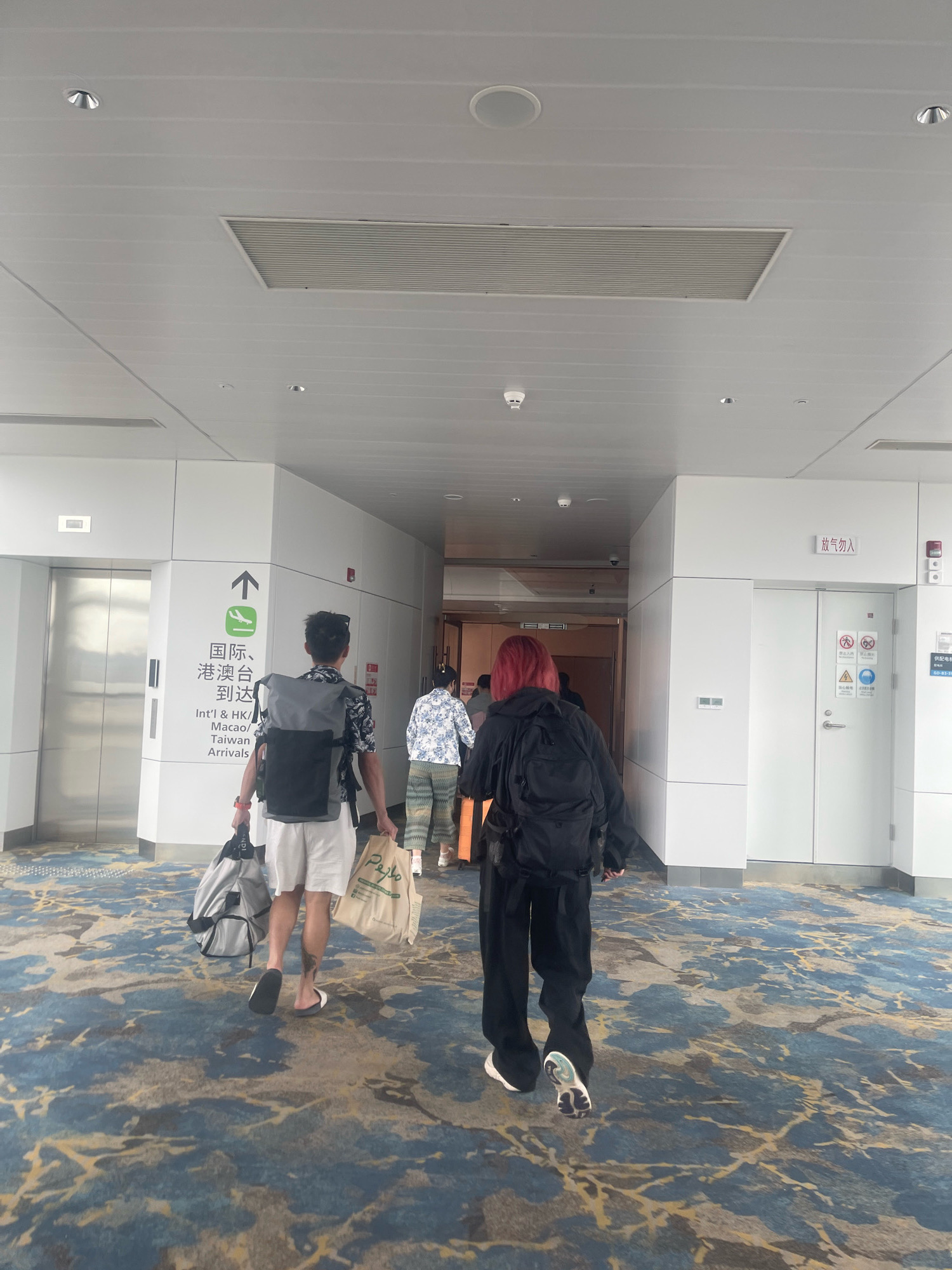 The image shows the interior of an airport terminal in Baiyun District, China, as evidenced by the Chinese signage. Two individuals are walking away from the camera down a wide, carpeted hallway with a patterned floor. The person in the foreground on the left is a male wearing a black and white floral short-sleeved shirt, white shorts, and black and white flip-flops, carrying a gray backpack on his shoulders and a tote bag in his right hand. The person to his right is a female with bright red hair, wearing a black hooded jacket with a backpack and black pants. Behind them, another person in a blue and white floral shirt and patterned pants is walking. The hallway features white walls, with an elevator door visible on the left and a set of double doors on the right. Overhead, there is a white ceiling with lighting fixtures and a ventilation system. Signs in both Chinese and English are visible, indicating "Int'l & HK/Macao/Taiwan Arrivals" near the elevator. The overall scene depicts travelers moving through an airport.