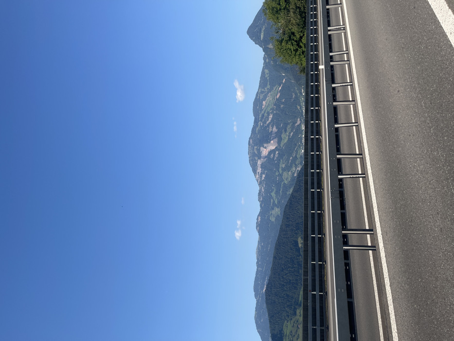 The image presents a static, elevated view of an asphalt highway in the vicinity of Buochs, Switzerland, under a clear, sunlit sky. In the immediate foreground, dark asphalt pavement is marked by two parallel, solid white lines. A grey metal guardrail, consisting of horizontal beams supported by vertical posts, extends horizontally across the lower half of the frame, serving as a barrier between the roadway and a drop-off or adjacent area. Beyond this, a secondary, darker grey concrete barrier with a lighter top rail is visible.
The midground and background are dominated by multiple ranges of mountains. These mountains are predominantly covered in dense green foliage and trees, with some higher peaks exhibiting visible light-colored rock faces. A small cluster of structures, indicative of a human settlement, is discernible at the base of the mountains in the distant left. The upper half of the frame displays an unobstructed expanse of bright blue sky, featuring two small, distinct white cumulus clouds and a single, minute dark object located in the upper left quadrant. The scene is devoid of human figures or active vehicles.