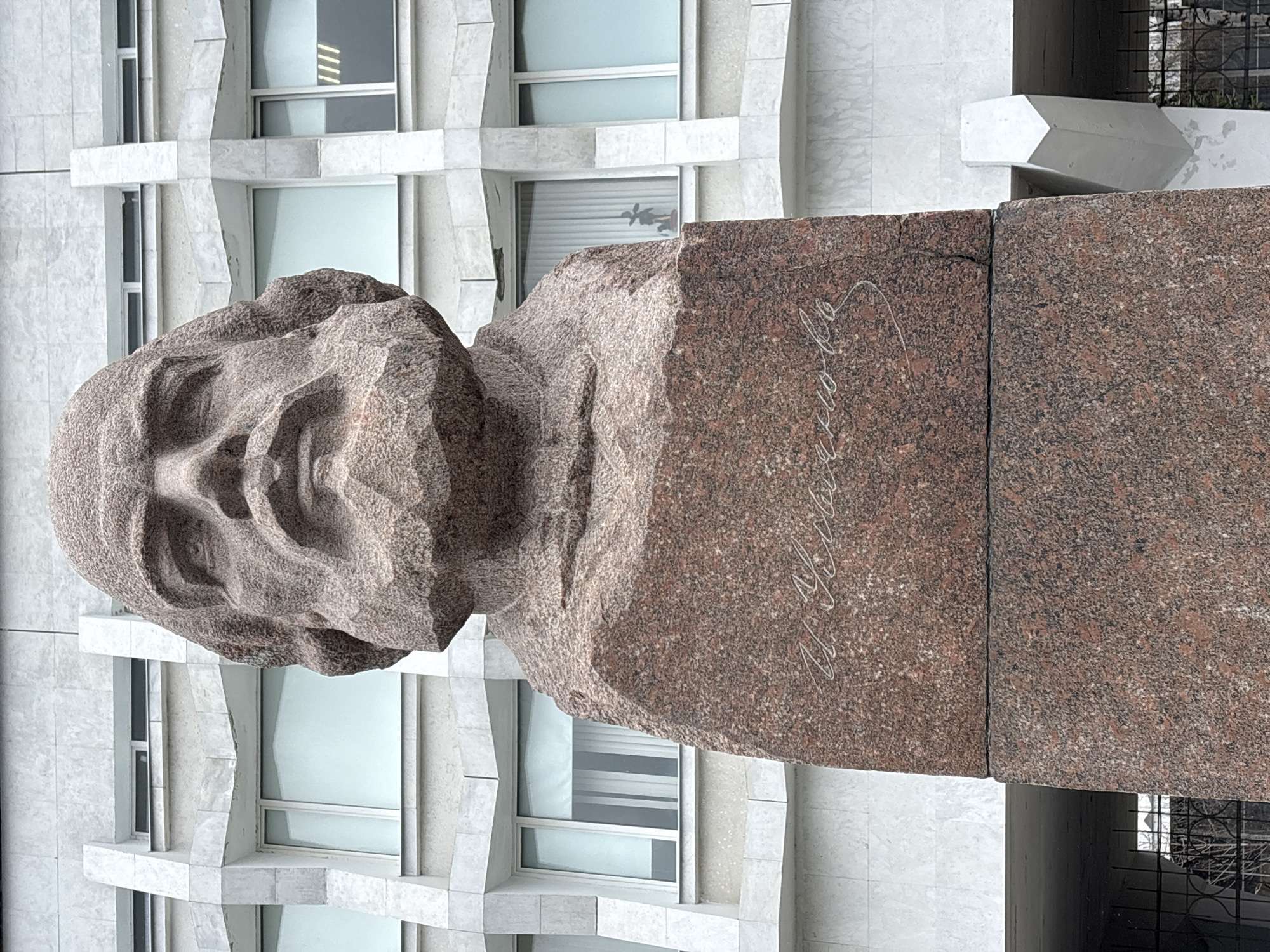 The image displays a large, imposing bust of a man, crafted from reddish-brown, speckled granite, prominently positioned outdoors. The bust depicts a male figure with a stern or contemplative expression, rugged facial features, and short, textured hair. It is mounted on a tall, rectangular pedestal, also made of the same coarse-grained stone.
On the front-facing side of the pedestal, a signature is carved in white cursive script, which clearly reads "В. Ульянов" (V. Ulyanov). This text, combined with the location context of Ulyanovsk, Russia, strongly suggests the monument commemorates Vladimir Ulyanov, famously known as Vladimir Lenin.
The bust and pedestal are set against the facade of a modern, multi-story building constructed from light-colored, possibly grey or white, stone or concrete panels. The building exhibits a geometric architectural style with rectangular windows framed in white. Some windows show reflections or interior blinds, and in one window, a small, dark, plant-like object is visible. A hint of a dark metal fence or grate can be seen in the lower right portion of the background.
The scene is illuminated by soft, diffuse daylight, indicating either an overcast sky or indirect light, as there are no harsh shadows. The overall impression is that of a public square or street in an urban environment, featuring a significant historical monument.
