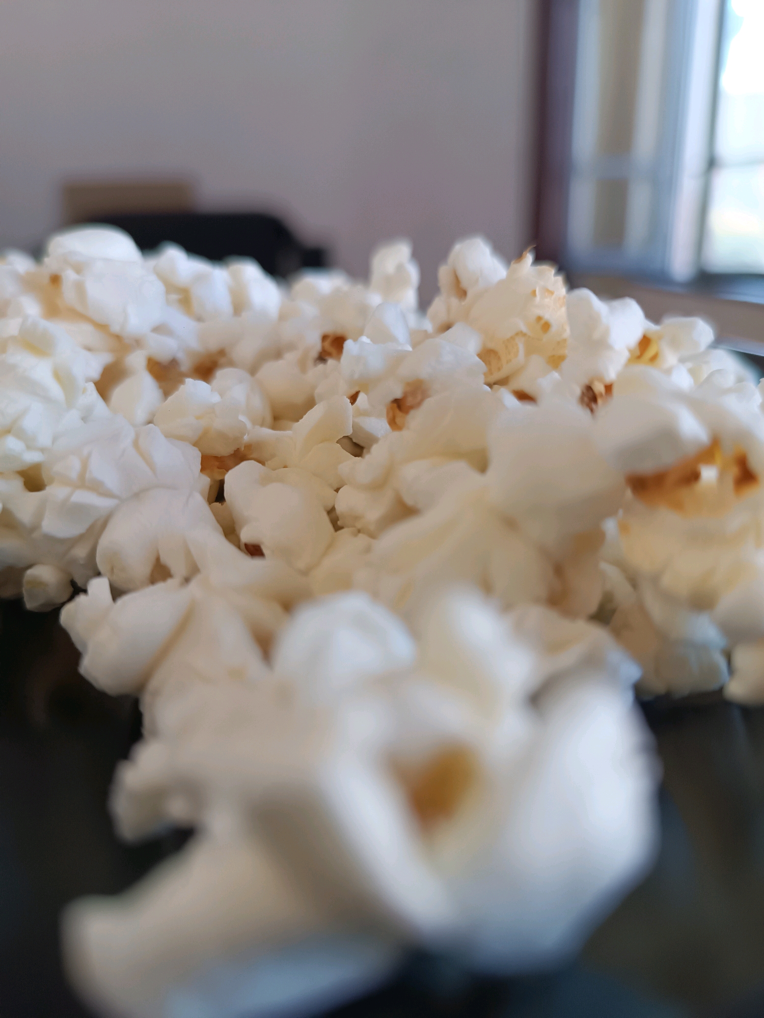 The image is a close-up, shallow depth of field shot of a pile of white popcorn. The popcorn is fluffy and irregular in shape, with some kernels showing brown, toasted parts. The foreground popcorn is blurred, while the popcorn in the midground is slightly more in focus, revealing more texture. The background is out of focus, showing a hint of a light-colored wall and a blurred window with a view of what appears to be trees and foliage. There are also dark shapes in the background that could be furniture. The lighting suggests an indoor setting with natural light coming from a window. There are no people, actions, or interactions depicted in the image. The location in Viadana, Italy, is not discernible from the image itself; this information must have been provided separately.