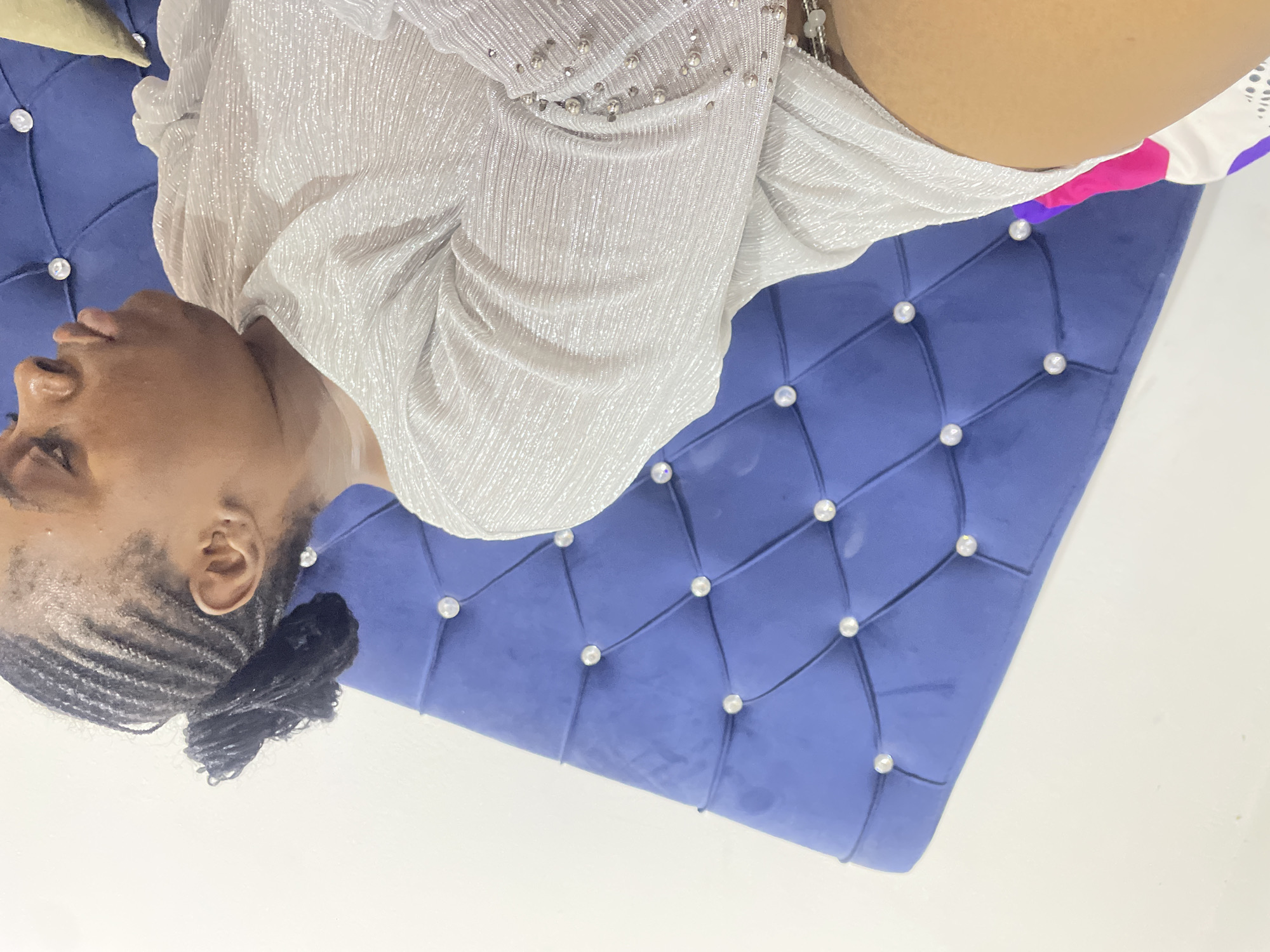 A woman with braided hair sits on a blue, button-tufted couch. She wears a light-colored, textured blouse. The scene appears to be indoors, possibly in a residential or commercial setting. The exact location in Abuja, Nigeria is not discernible from the image.