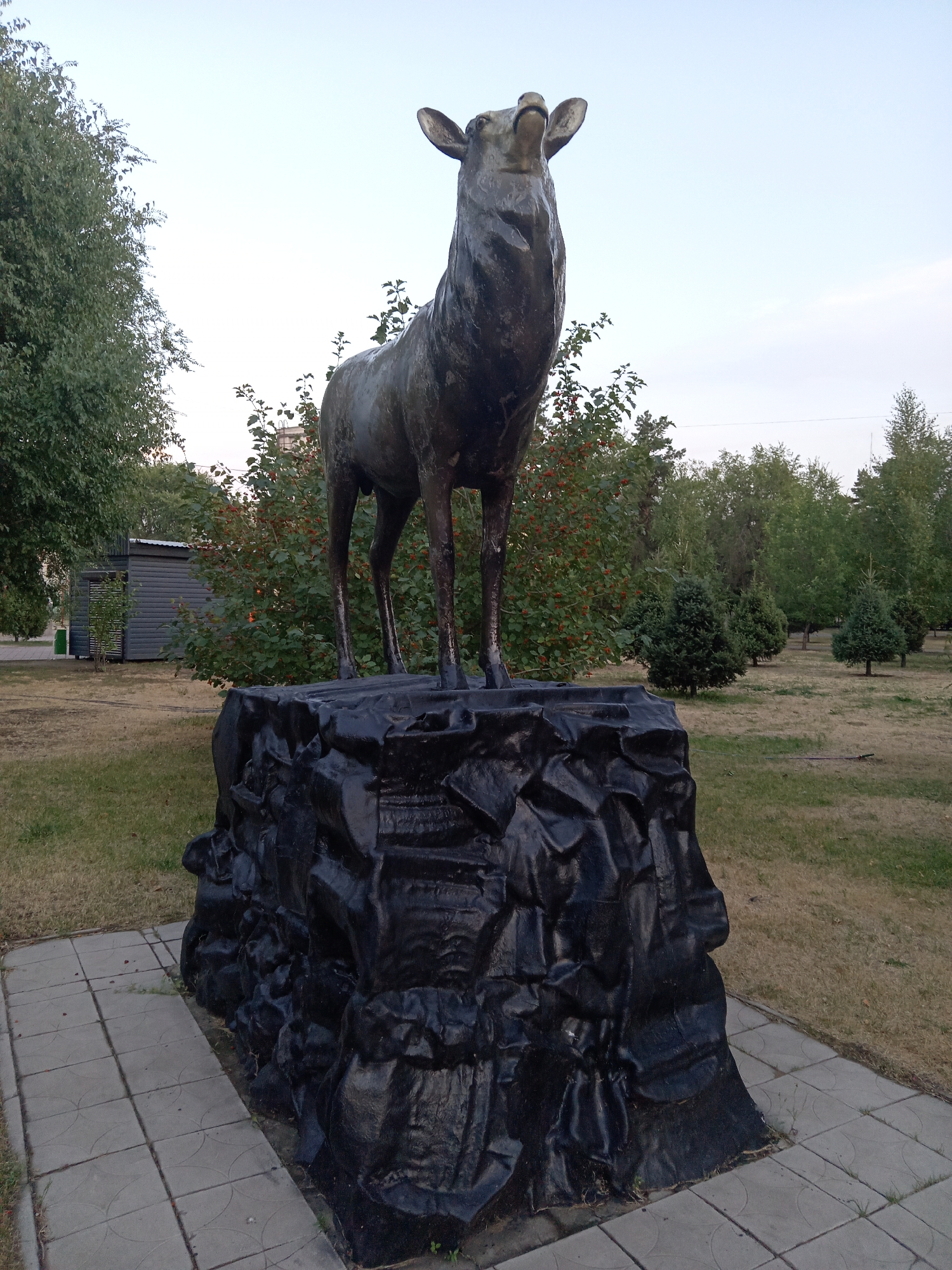 A metal sculpture of a deer stands on a jagged, dark, rock-like pedestal in a park in Rudny, Kazakhstan. The deer is bronzed and facing upwards with its head tilted back and mouth open. The pedestal is irregularly shaped and appears to be made of painted metal or concrete, with a rough, textured surface. The deer is positioned on a paved area made of square concrete tiles with a decorative pattern. Behind the sculpture, there is a lawn, trees with green and red foliage, and a small grey building with a slanted roof. The sky is overcast with a pale blue hue. The overall setting suggests an outdoor public space in a city.
