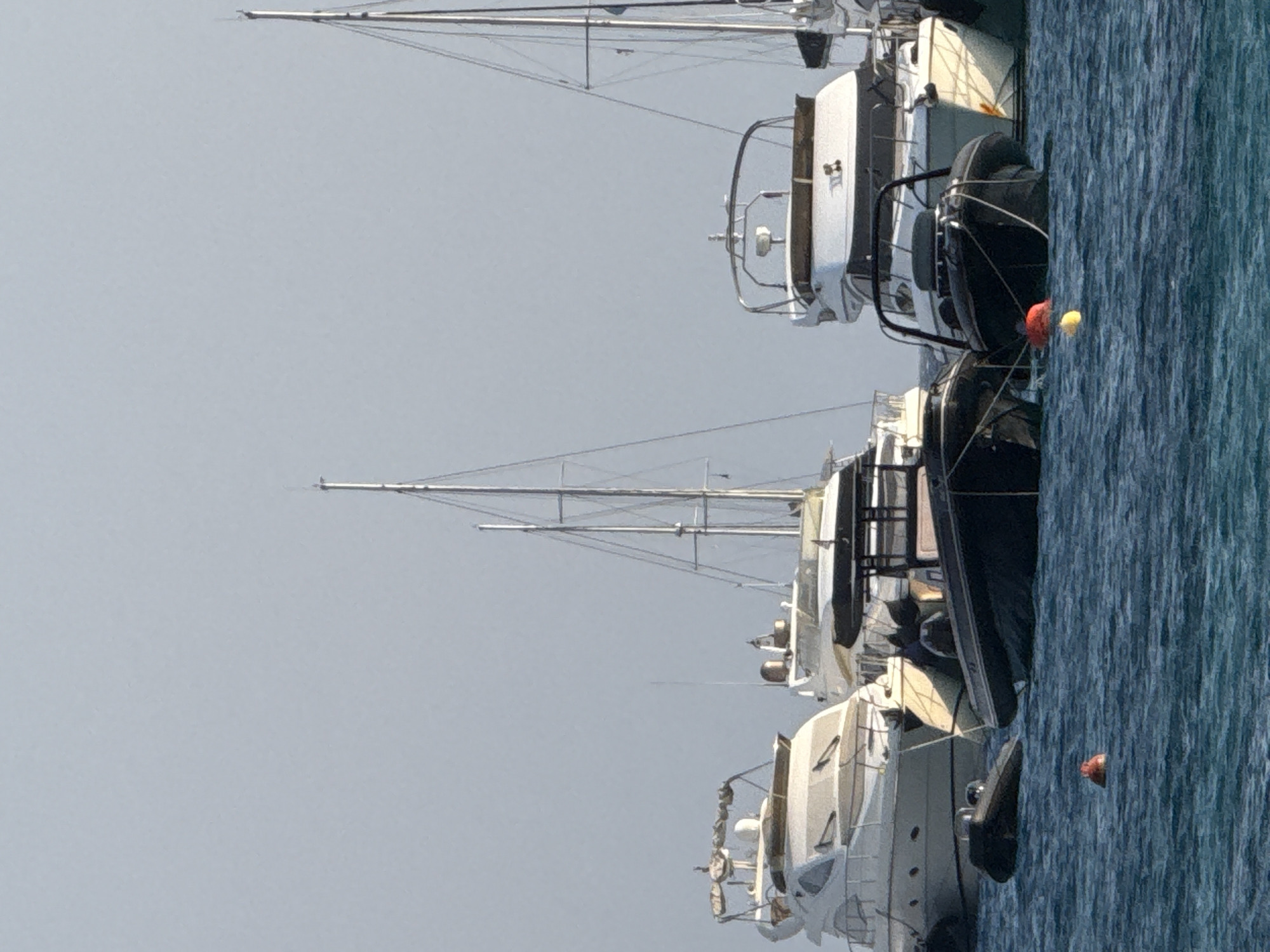 In the waters of Agios Ioannis Diakoftis, Greece, several vessels are anchored. In the foreground, two rigid inflatable boats (RIBS) are prominently positioned, one black and one dark grey. Both are secured with ropes, and the black RIB has a yellow and red buoy nearby in the turquoise water. Behind them, a large white yacht with multiple decks is visible, appearing to be a motor yacht. Further back, another white yacht, also a motor yacht, is partially obscured. Tall masts of a sailing vessel rise above the motor yachts on the right side of the frame. The sky is a pale, hazy blue, suggesting a clear day with minimal cloud cover. The water is moderately choppy. No people are clearly discernible in the image.