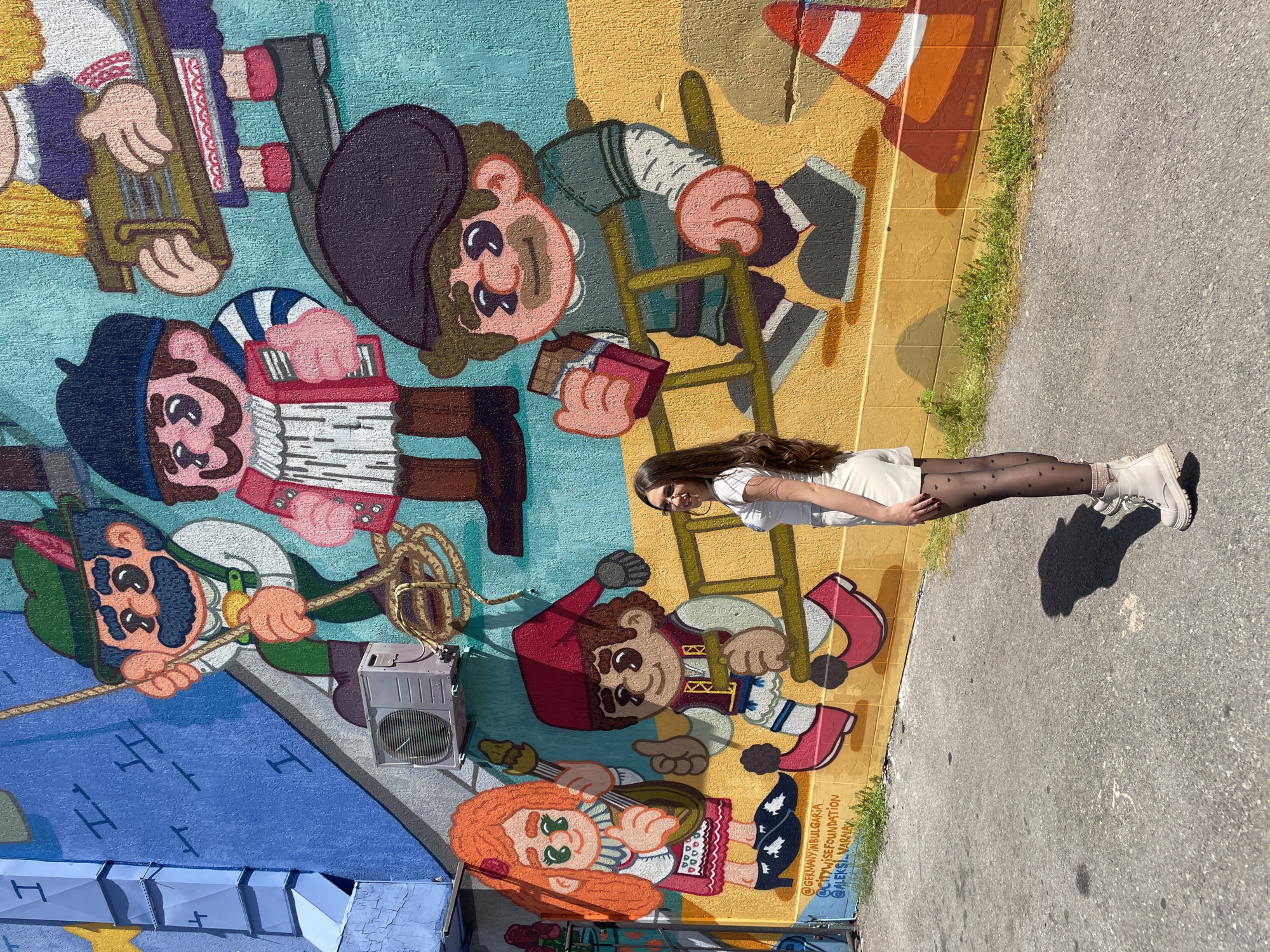A young woman poses in front of a large mural in Sofia, Bulgaria. The mural depicts several cartoon-like characters with Slavic features, playing musical instruments and engaging in activities like climbing a ladder. The mural is painted on a wall with sections colored in blue, yellow, and orange, and features an air conditioning unit mounted on the wall. The woman is wearing a light-colored dress, patterned tights, and boots. The ground is paved and shows her shadow. The lower right corner of the mural includes the text "@GERMANYINBULGARIA @CITYWISEFOUNDATION @ALEKSII.VARAKIN."