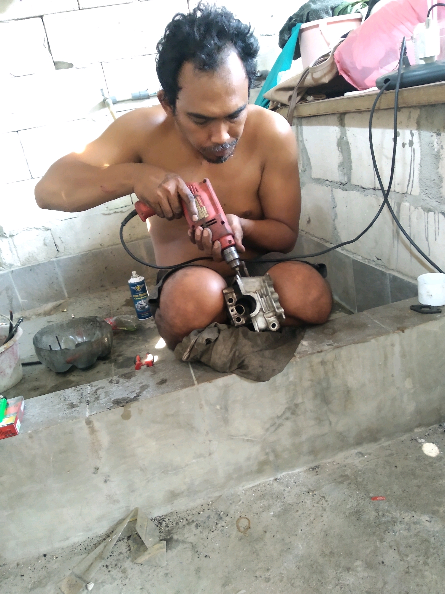 A shirtless adult male with dark hair is seated, facing away from the camera and to the left, as he operates a red power drill on a piece of machinery. The man is positioned within a large, rectangular concrete structure that appears to be a water basin or cistern. The surrounding environment is industrial or utilitarian, with unpainted cinder block walls and a concrete floor covered in dust and debris.
The man is holding the drill with both hands, directing its tip into the component. He is wearing dark shorts. To his left, on the floor of the concrete basin, are several items including a partially cut plastic bottle, a can of spray lubricant, and a piece of dark cloth on which the machinery rests. A second, smaller red object is on the floor near the spray can.
Behind the man, resting on a ledge or shelf against the cinder block wall, are several objects. These include what appears to be a pink duffel bag or large piece of fabric, a beige bag, and some green fabric. A black electrical cord from the drill extends upwards and to the right.
The overall scene suggests an informal workshop or maintenance area, possibly in a residential or small industrial setting within South Tangerang, Indonesia. The lighting is bright, suggesting daylight, with some shadows indicating overhead illumination. The concrete basin is wet in places, with visible water marks on the walls.