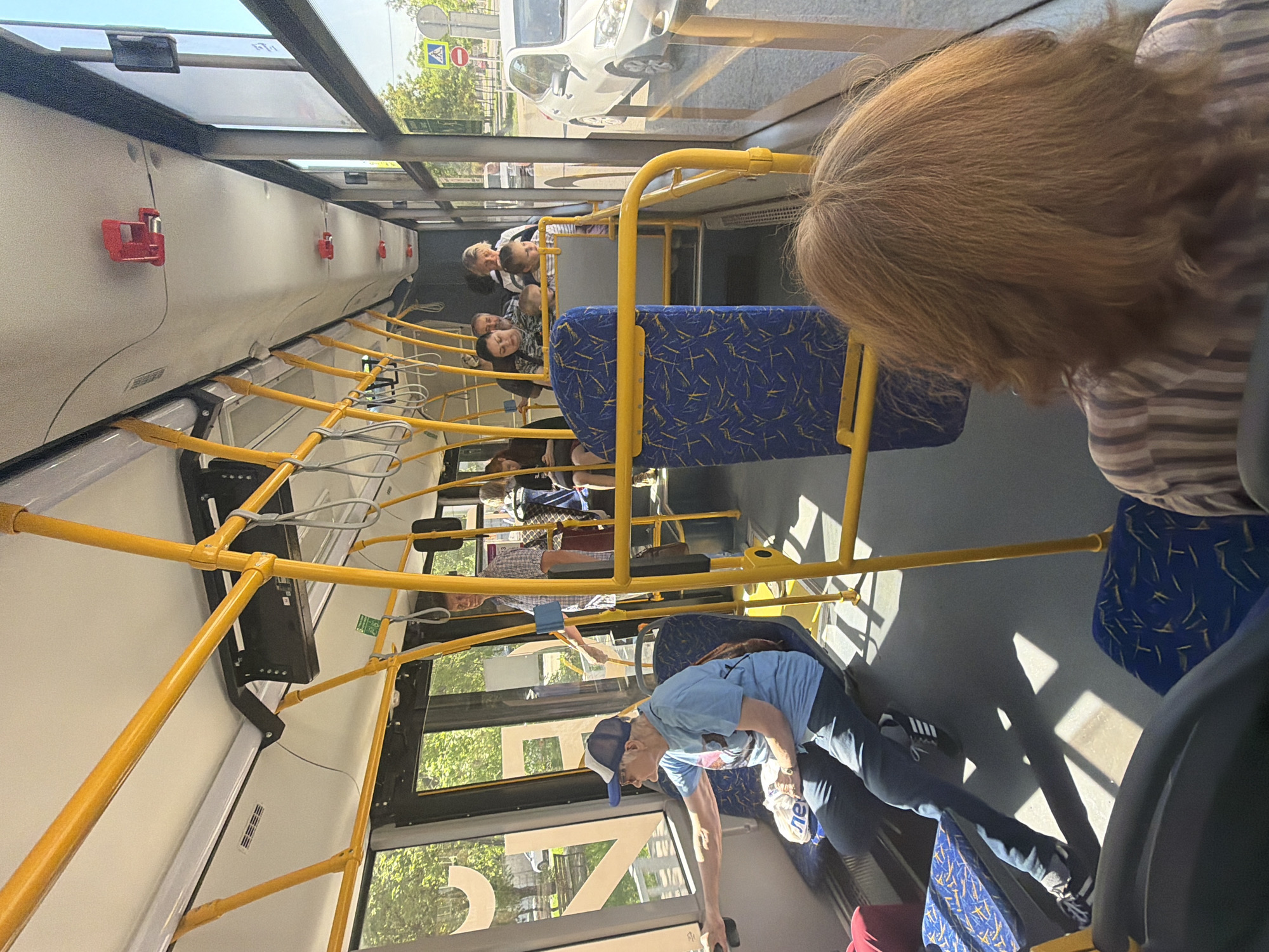 The image captures the interior of a public bus on a bright, sunny day, likely in Krasnoyarsk, Russia. The bus is occupied by several passengers, with strong sunlight streaming through the windows and casting sharp shadows across the floor and seats.
In the immediate foreground, seen from behind, is a person with reddish-brown hair, wearing a striped top, possibly seated or standing. Further into the bus, on the right side, an older man wearing a blue cap, blue t-shirt, and dark pants is leaning forward, holding a white plastic bag with "НЕ" visible on it. He appears to be attending to something or placing an item. Beside him, a blue patterned seat is visible.
On the left side of the bus, a group of passengers is seated, including a blonde-haired woman and other individuals, some appearing to be children or younger adults. They are looking forward or conversing. Further back on the left, a man in a plaid shirt stands, holding onto one of the many yellow grab poles that crisscross the bus interior.
The bus features large windows, through which a white car, trees, and street signs (including a pedestrian crossing sign) are visible, indicating an urban or suburban setting. The interior walls are light-colored, fitted with several red emergency hammers or buttons, and a black digital display screen. The bright weather suggests it is daytime. A large reflection resembling "27" is visible on the window near the leaning man. Overall, the scene depicts a typical public transport environment during a sunny period.