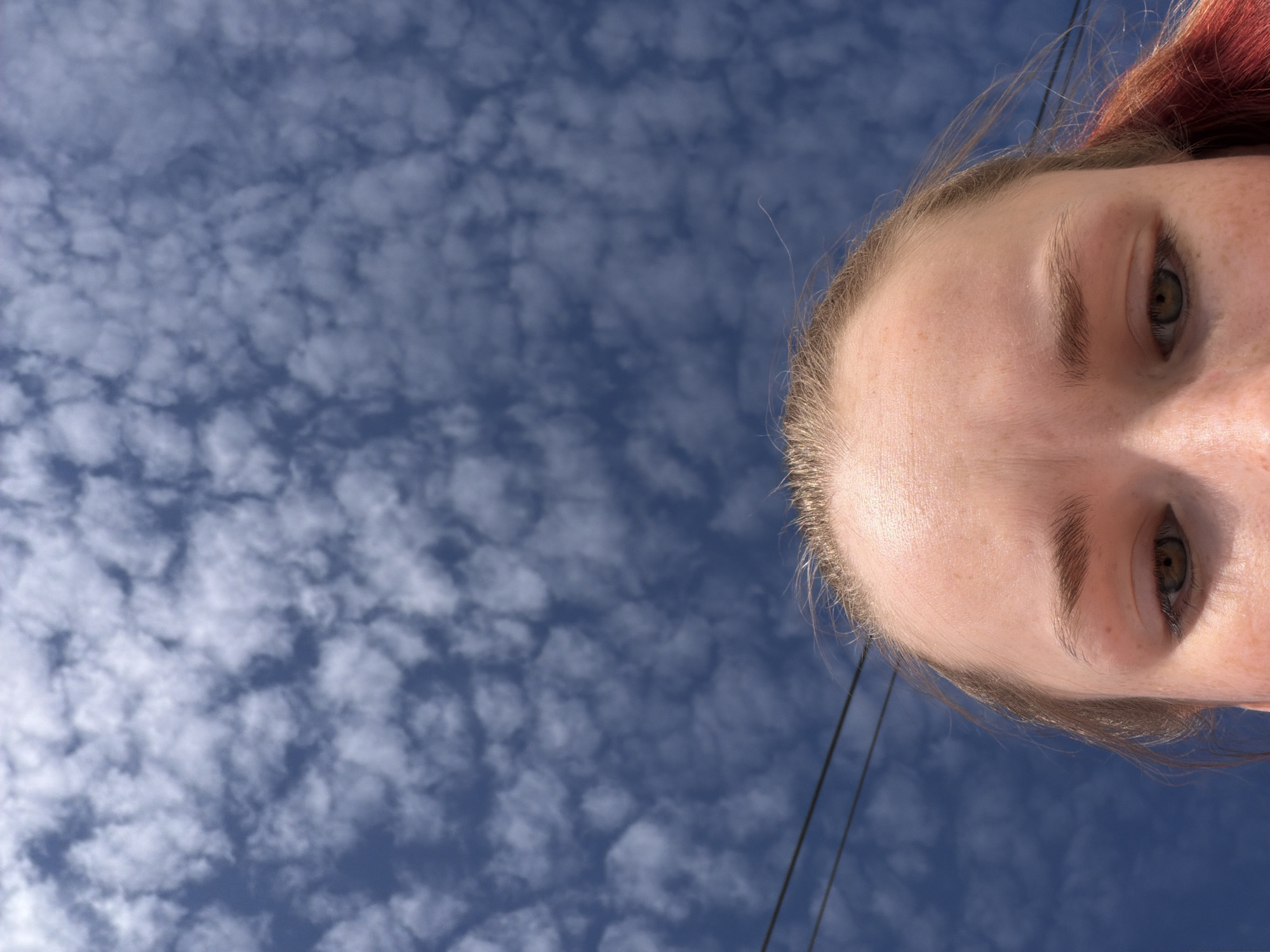 The image depicts a view of a blue sky dotted with small, white clouds, with the face of a fair-skinned person in the lower right portion of the frame. The person has reddish-brown hair near the top of the head and visible eyebrows, eyes, and forehead. Two thin, dark lines, possibly electrical wires, stretch from the top of the image down towards the person's face. No specific details indicate the location is Berezniki, Russia.