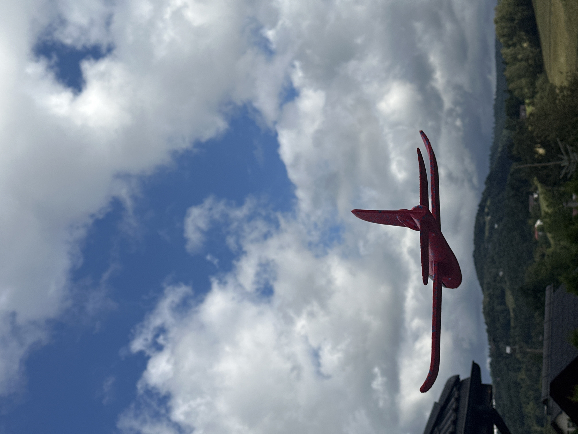An overhead low-angle shot shows a red toy airplane against a backdrop of a cloudy blue sky and a wooded, hilly landscape. The toy airplane is positioned in the center of the frame, with its wings extended and its body facing right. The sky is a mixture of blue and white clouds, with some darker, heavier clouds towards the bottom of the frame. Below the sky, there is a green, hilly landscape dotted with trees and some houses, suggesting a rural or suburban setting. A dark-colored, sloped roof structure is visible on the left side of the image, partially obscuring the landscape. The overall scene appears to be outdoors, possibly on a cloudy day.