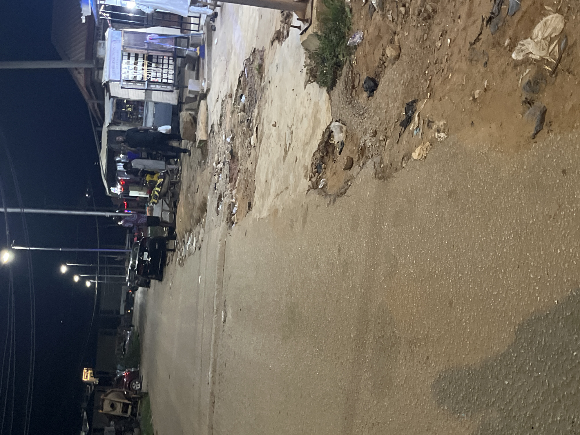 The image captures a street scene at night in Abuja, Nigeria, characterized by a mix of parked vehicles, makeshift stalls, and uneven road conditions. The street is paved with asphalt and exhibits significant deterioration, with broken sections and exposed earth on the right side of the road. Several cars, including a black sedan and what appears to be a red SUV, are visible. People are present, with a few individuals standing near stalls and others near vehicles. One person in a dark outfit is holding a white bag. The stalls appear to be small, open-fronted structures, with one displaying the words "CHARGING POINTS." Streetlights illuminate the scene, casting shadows and highlighting the debris scattered along the roadside. The environment appears to be a commercial or residential area with limited infrastructure.
