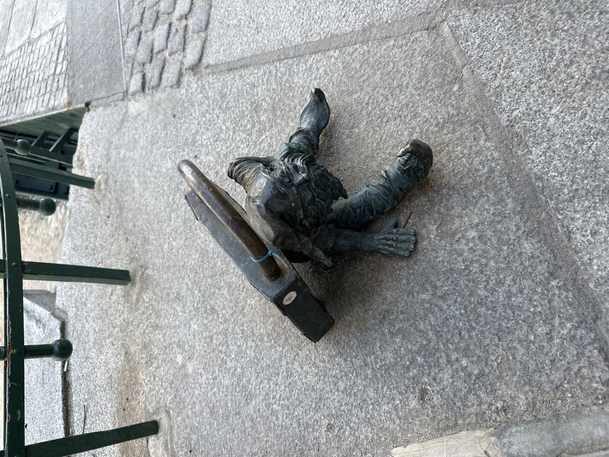 The image depicts a bronze dwarf statue lying prone on a stone surface in Wrocław, Poland. The dwarf is positioned as though he has fallen forward, his limbs splayed out. He wears an outfit common to the Wrocław dwarf statues. He carries a flat, rectangular object. Part of a dark green metal railing is visible near the left edge of the frame. The stone surface is textured and appears to be a sidewalk or plaza area. The lighting is bright, suggesting an outdoor daytime setting.