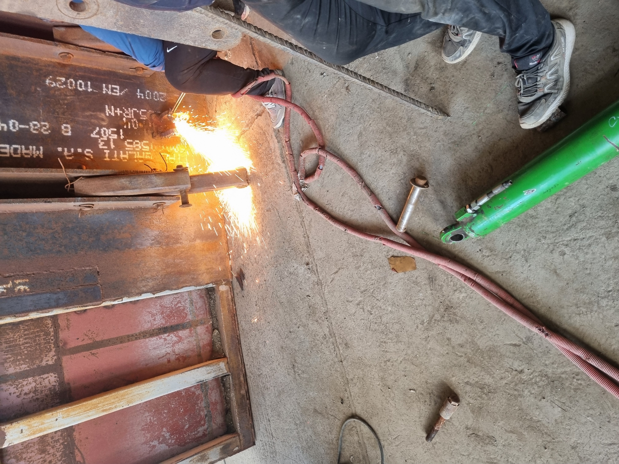 An overhead shot depicts a worker torch-cutting a metal structure, generating sparks. The worker is wearing dark pants, a blue shirt, and grey athletic shoes. A twisted, red hose stretches from the torch to a green hydraulic cylinder resting on the concrete floor. Debris is scattered around the workspace. Text "MELATI S.A. MADE" is visible on the structure. The location is unidentified, with no visible clues indicating Ovcharovo, Bulgaria.