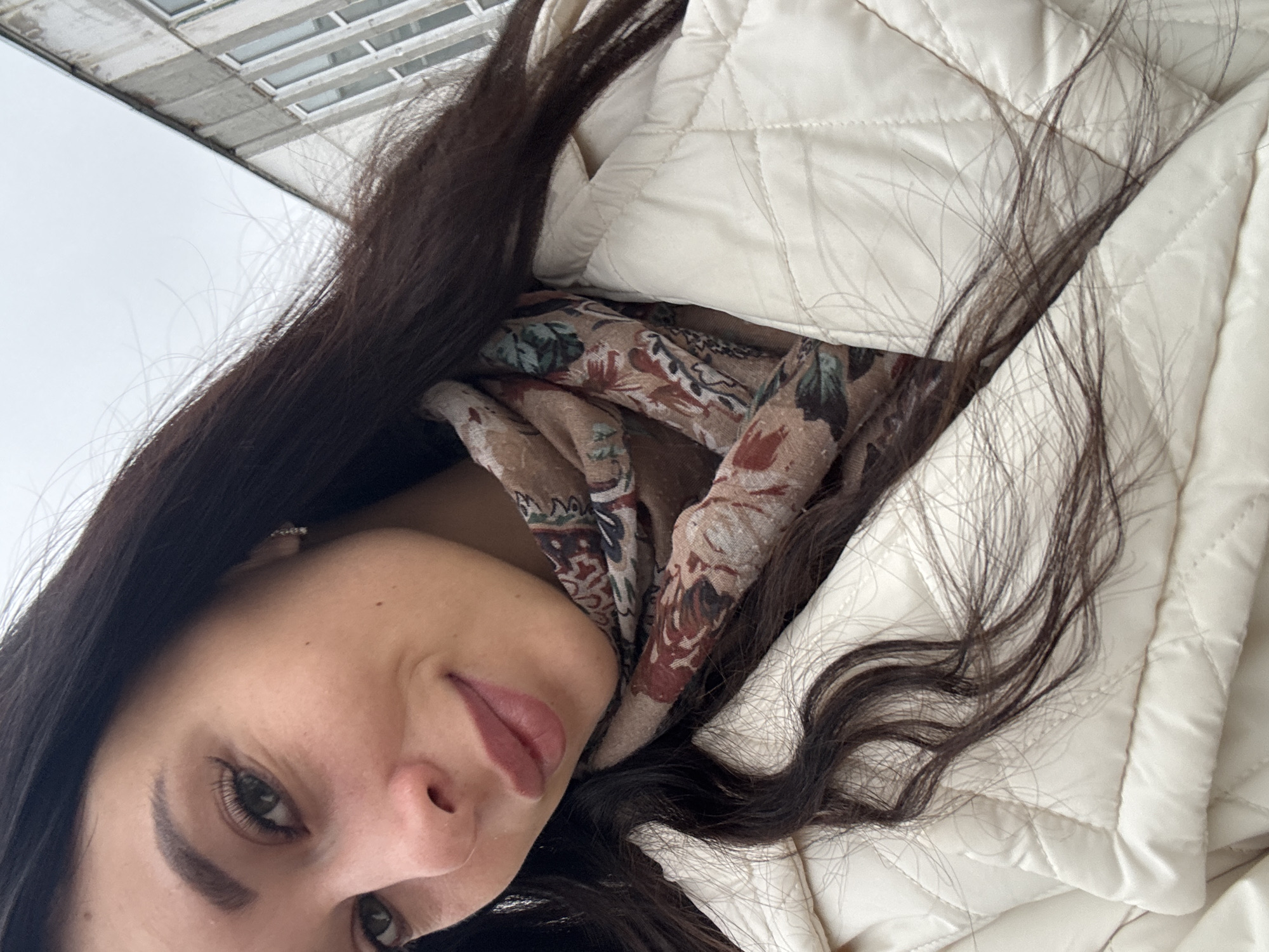 A woman with dark hair is pictured in what appears to be a selfie. She is wearing a white quilted jacket and a patterned scarf. Outside, in the background, a multistory building with visible windows is present. Given the request for location detail, it's speculated the image was taken in an urban setting in Chaikovsky, Russia, based on the user's location. However, this identification is based solely on user-provided information, as no specific locational details can be definitively determined from the image itself.