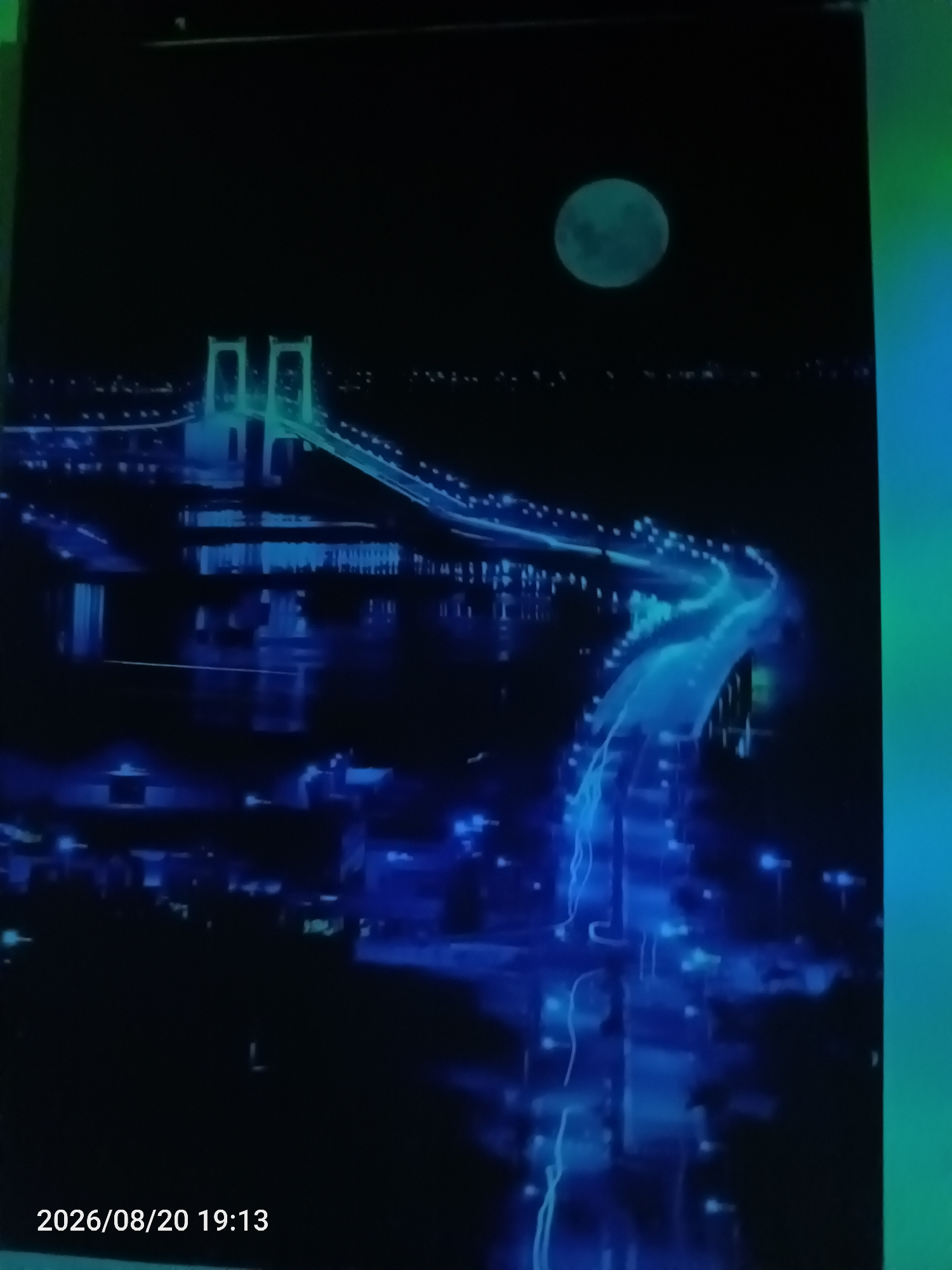 The image depicts a nighttime cityscape illuminated by bright blue and green lights, with a prominent bridge structure and a large, pale circle in the dark sky, likely the moon. The bridge appears to be suspended over water, with glowing lights tracing its path and extending onto a road. The surrounding area shows clusters of smaller lights, suggesting buildings and other urban infrastructure. The overall impression is one of a vibrant, modern city at night, possibly with futuristic elements due to the intense, stylized lighting. The text overlay at the bottom left indicates a date and time: "2026/08/20 19:13". The right side of the image shows a gradient of green and blue light, suggesting an artificial light source or effect. There are no identifiable people or specific actions depicted in the scene. The location is described as Jawilan, Indonesia, however, the visual elements do not provide specific landmarks to confirm this location.
