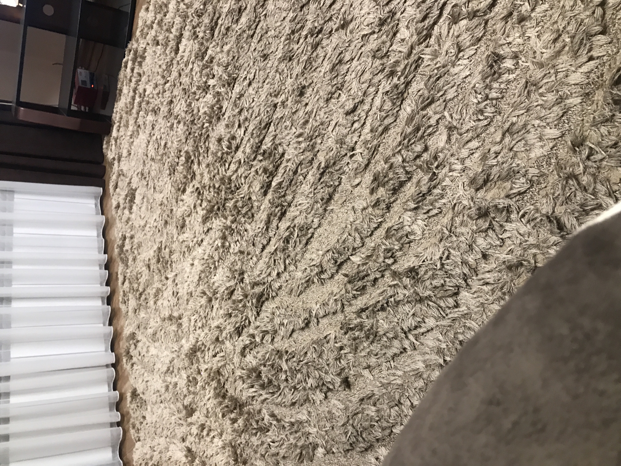The image shows a close-up view of a shag rug with a neutral color palette. To the left, there are white blinds. Above the blinds is part of a brown-toned entertainment center. A portion of a gray-colored object, possibly a piece of furniture, appears in the bottom right of the image. The location cannot be ascertained from the visual information provided.